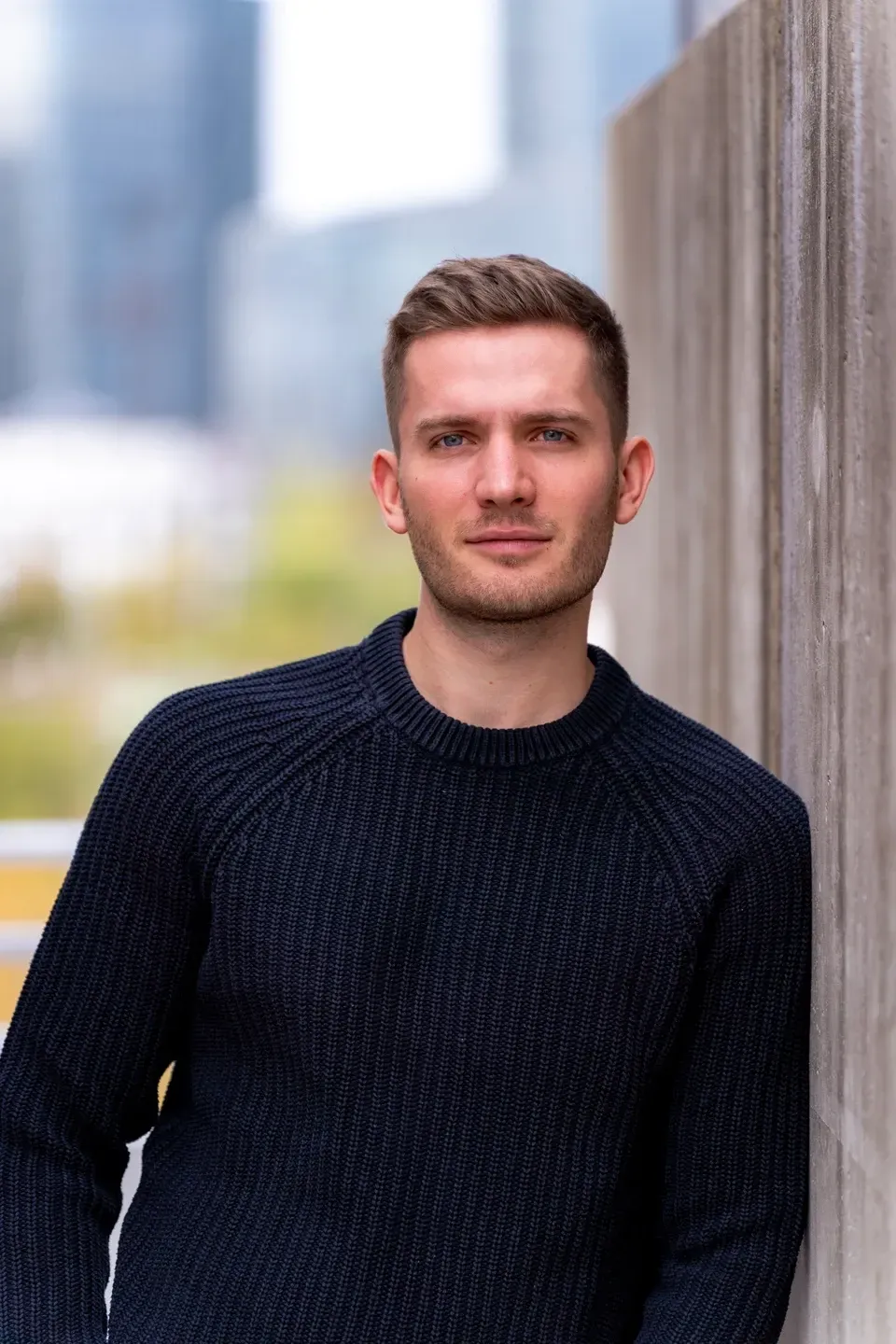 Mann im dunkelblauen Strickpullover lehnt an einer Betonwand, im unscharfen Hintergrund Hochhäuser