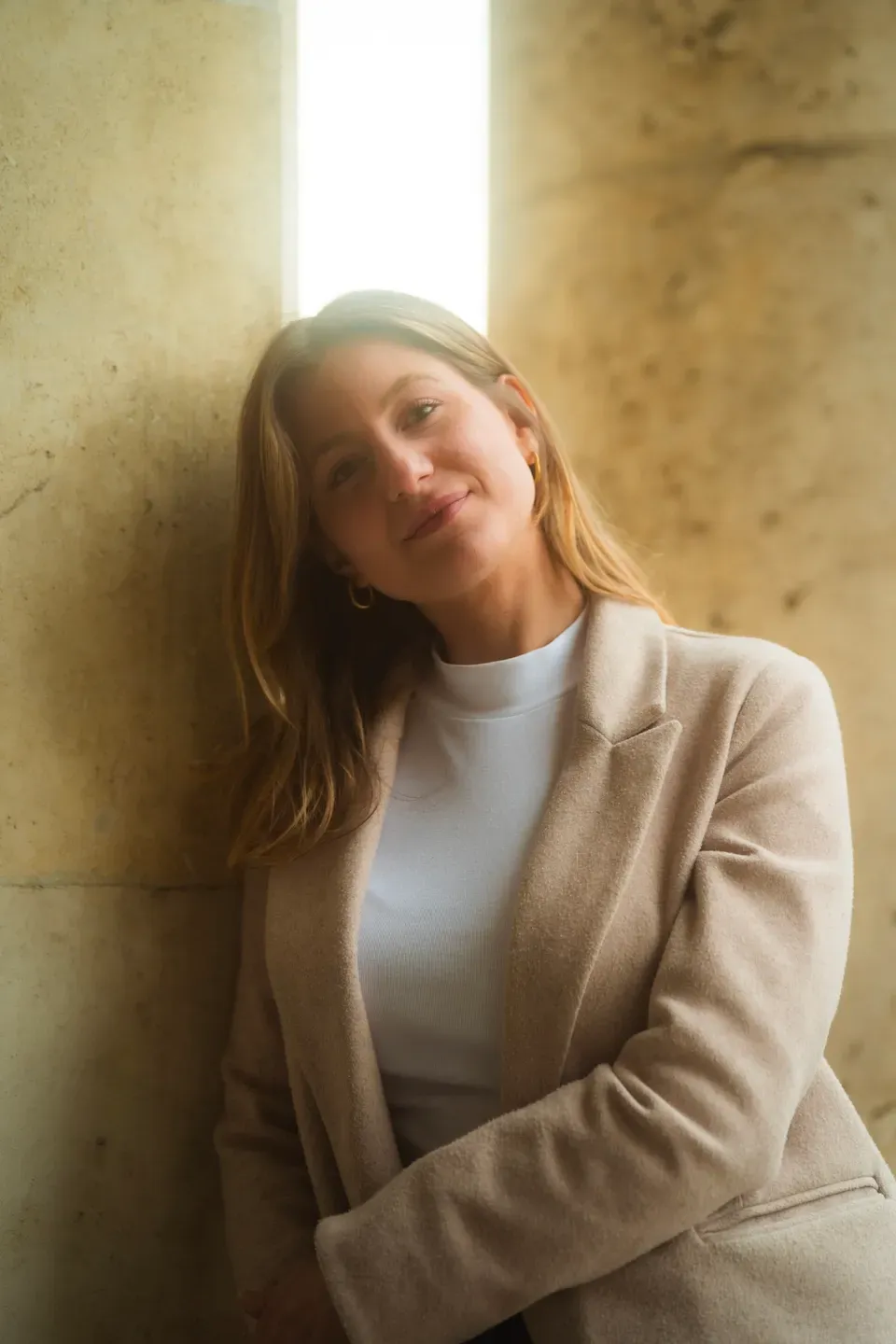 Frau im beigen Blazer lehnt lächelnd an einer Steinwand, hinter ihr fällt Licht durch einen schmalen Spalt