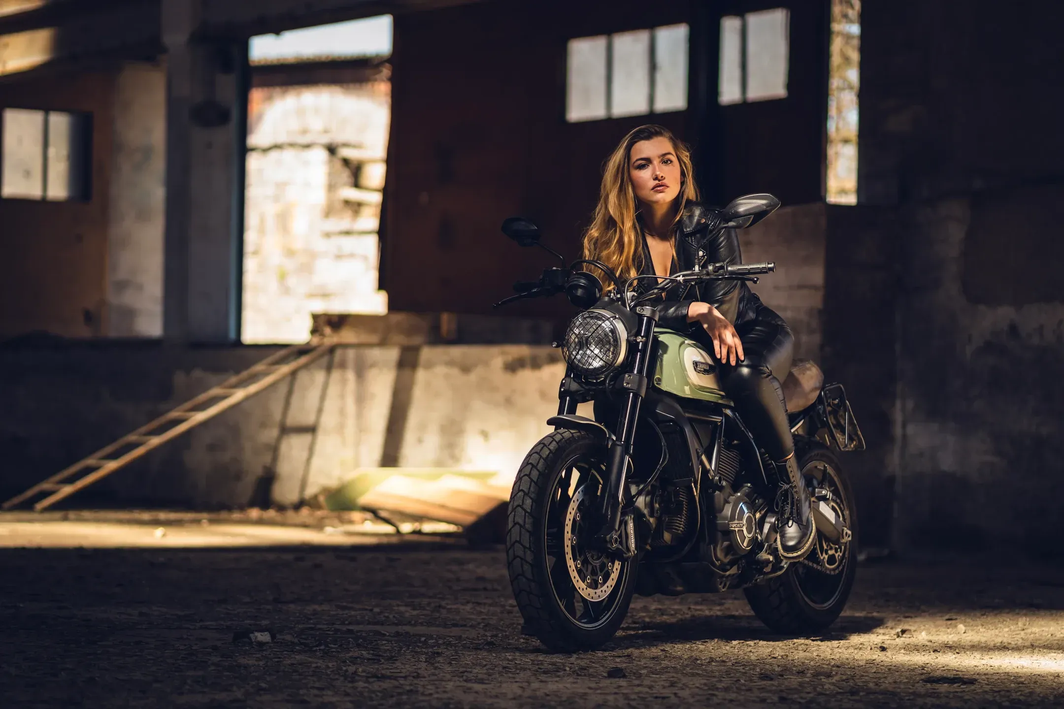Frau in Lederjacke sitzt auf einem Scrambler-Motorrad in einer verlassenen Halle, seitliches Sonnenlicht