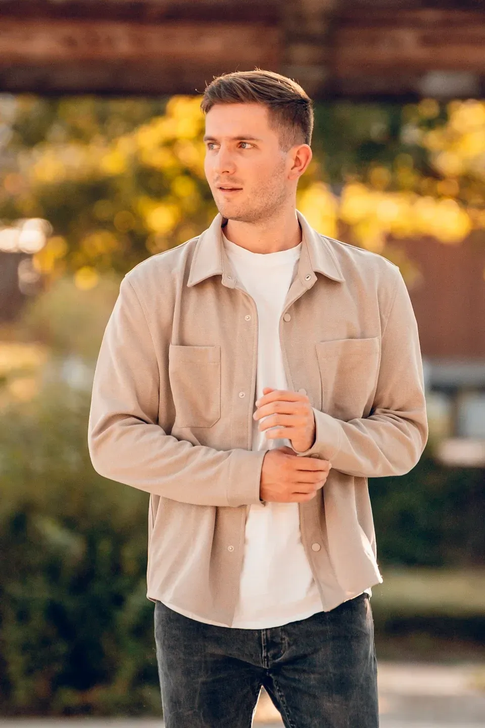 Mann in beiger Hemdjacke und weißem Shirt steht im Freien und blickt zur Seite, dahinter gelbes Laub