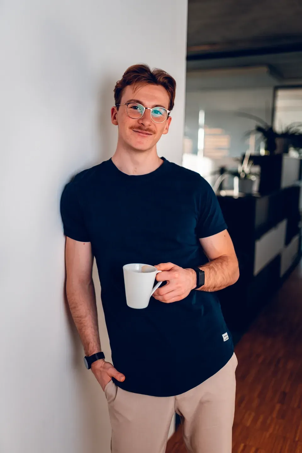 Junger Mann mit Brille und dunklem T-Shirt lehnt an einer weißen Wand im Büro und hält lächelnd eine Kaffeetasse