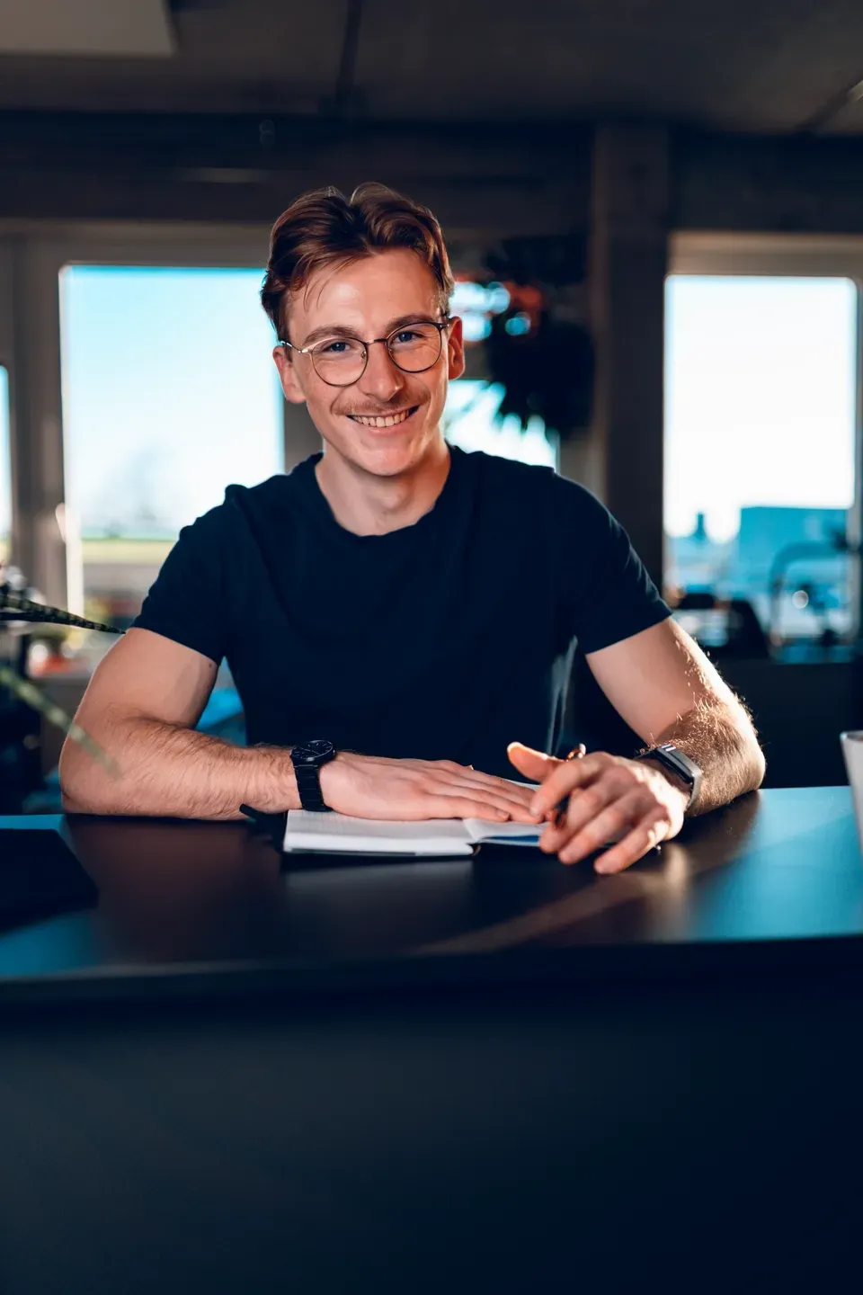 Junger Mann mit Brille sitzt lächelnd am Schreibtisch vor großen Fenstern, die Hände auf einem Notizbuch