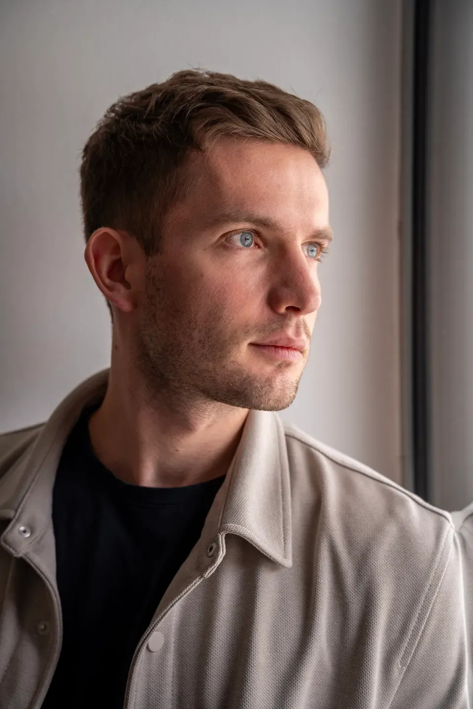 Mann mit kurzen Haaren und heller Jacke blickt nachdenklich zur Seite aus dem Fenster, weiches Tageslicht