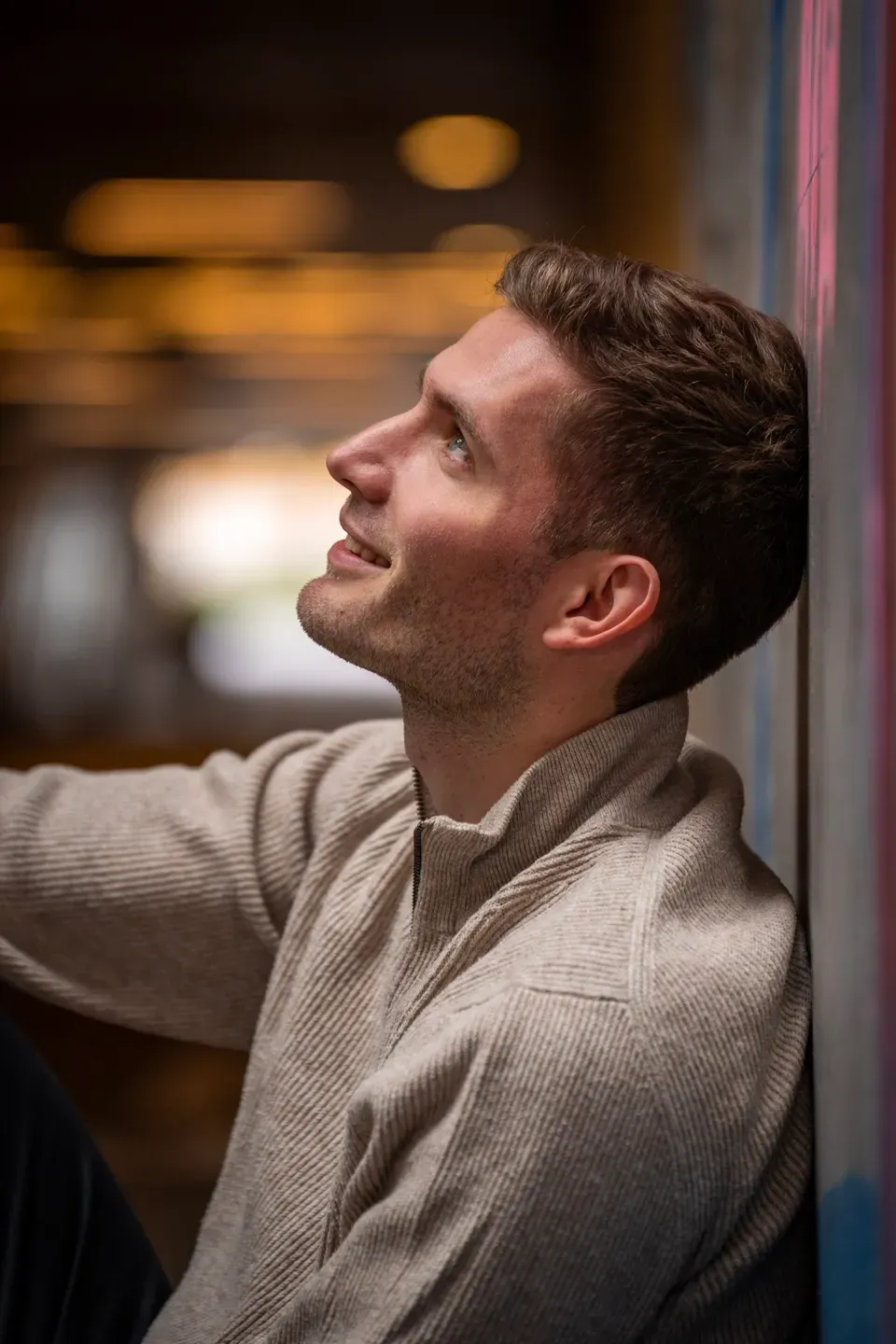 Mann im beigen Strickpullover lehnt an einer Wand und blickt lächelnd nach oben, Hintergrund in warmem Bokeh
