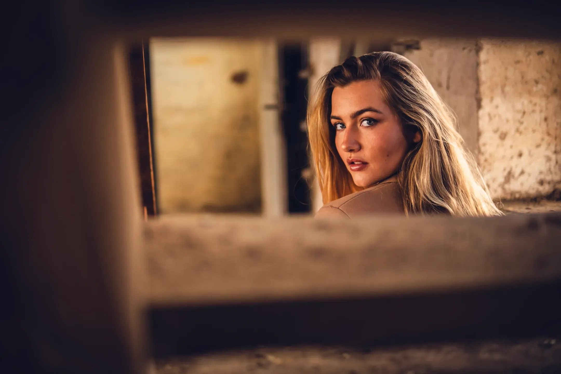 Blonde Frau blickt über die Schulter durch eine Öffnung in einer alten Steinmauer, warmes Licht im Gemäuer