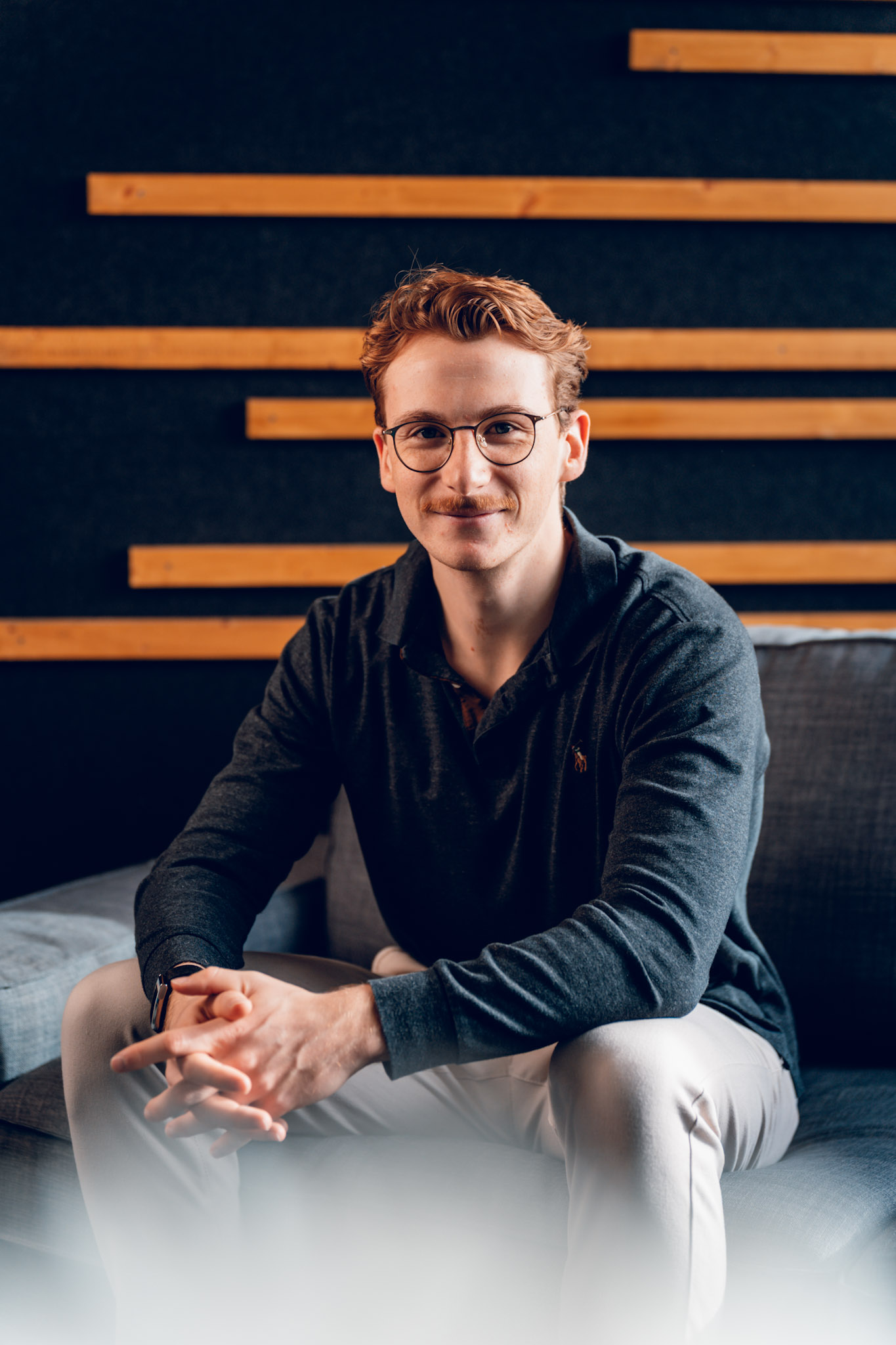 Junger Mann mit Brille und dunklem Pullover sitzt lächelnd auf einem Sofa vor einer Wand mit Holzleisten