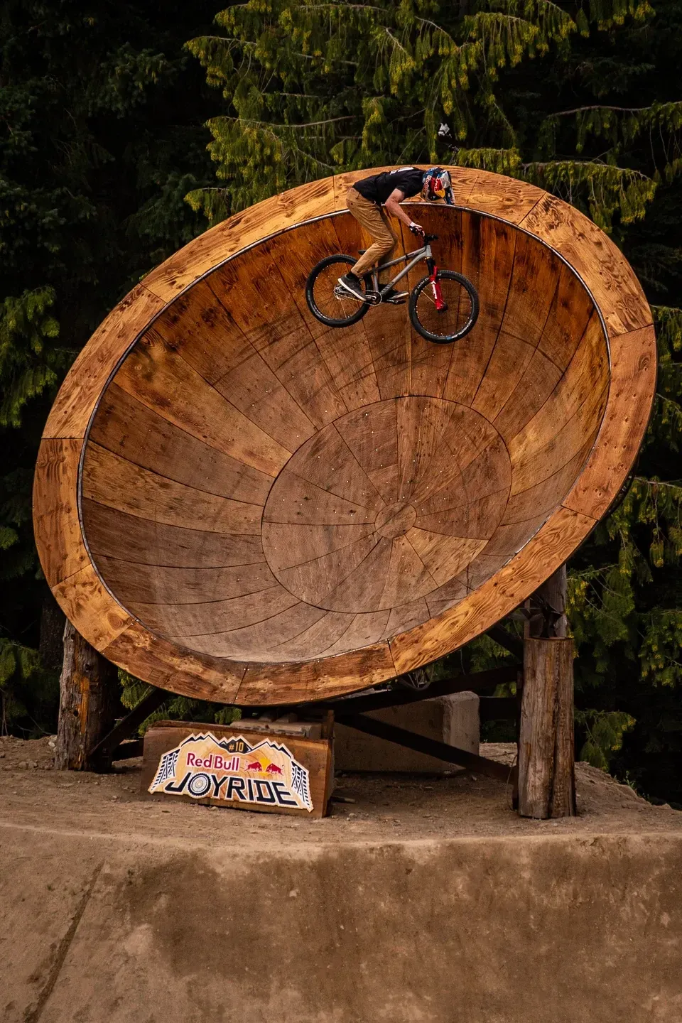 Mountainbiker fährt am Rand einer riesigen Holzschüssel, darunter ein Red Bull Joyride Schild, dahinter Nadelwald