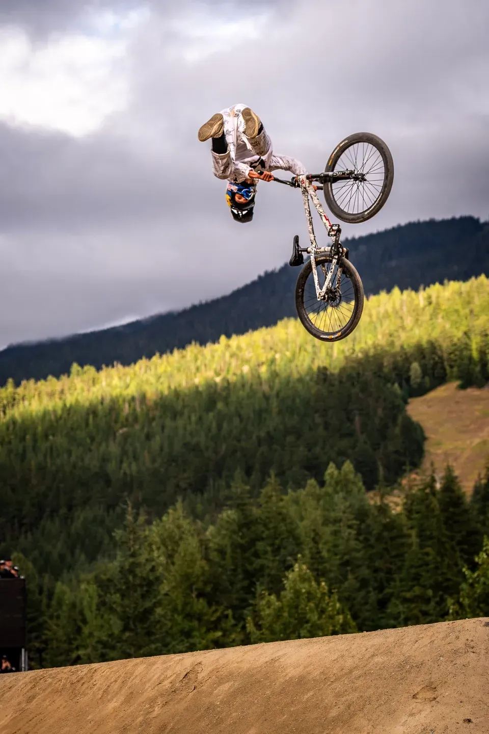 Mountainbiker kopfüber im Backflip über einer Erdrampe, bewaldeter Berghang unter grauen Wolken