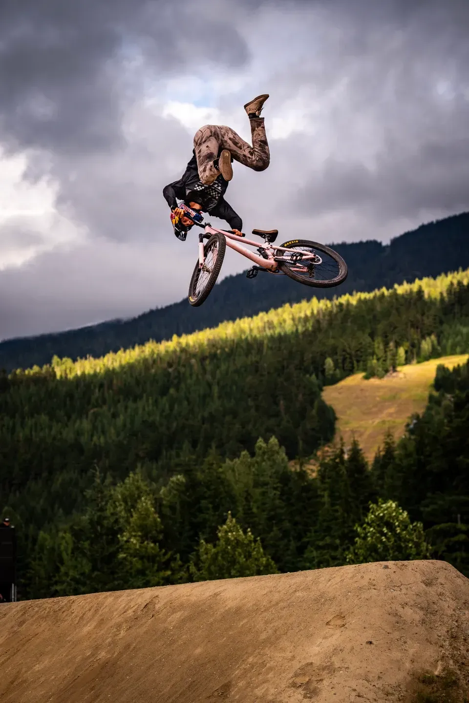 Mountainbiker mit rosa Rad im Flip kopfüber in der Luft, sonnenbeschienener Waldhang und dunkle Wolken