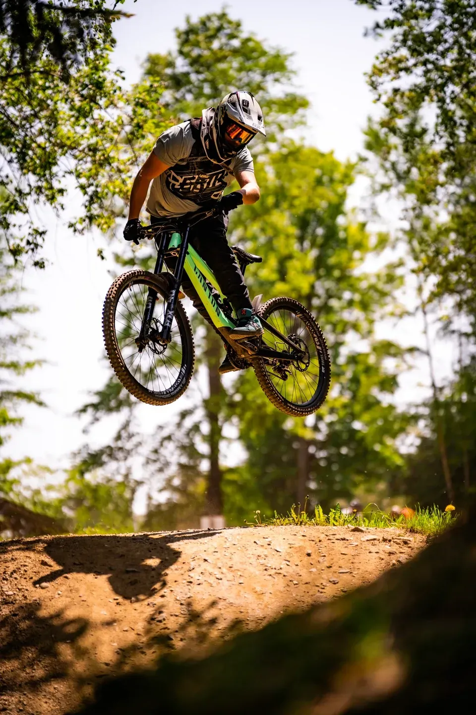 Mountainbiker mit Integralhelm springt über eine Erdkante im sonnigen Laubwald, Schatten auf dem Boden