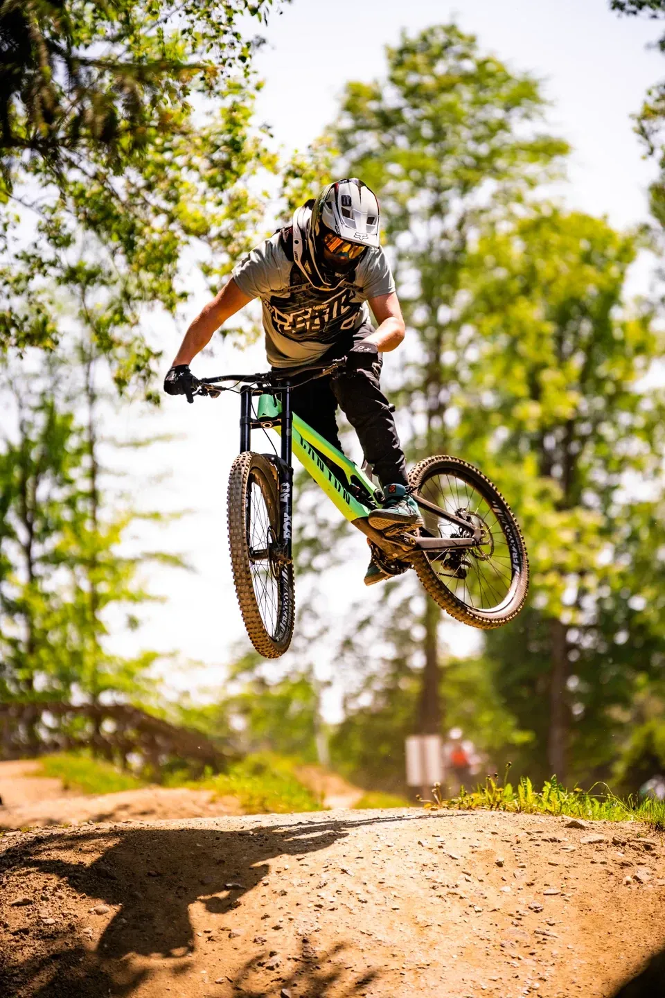 Mountainbiker mit grünem Rad springt frontal auf die Kamera zu, Untersicht vor hellen Laubbäumen