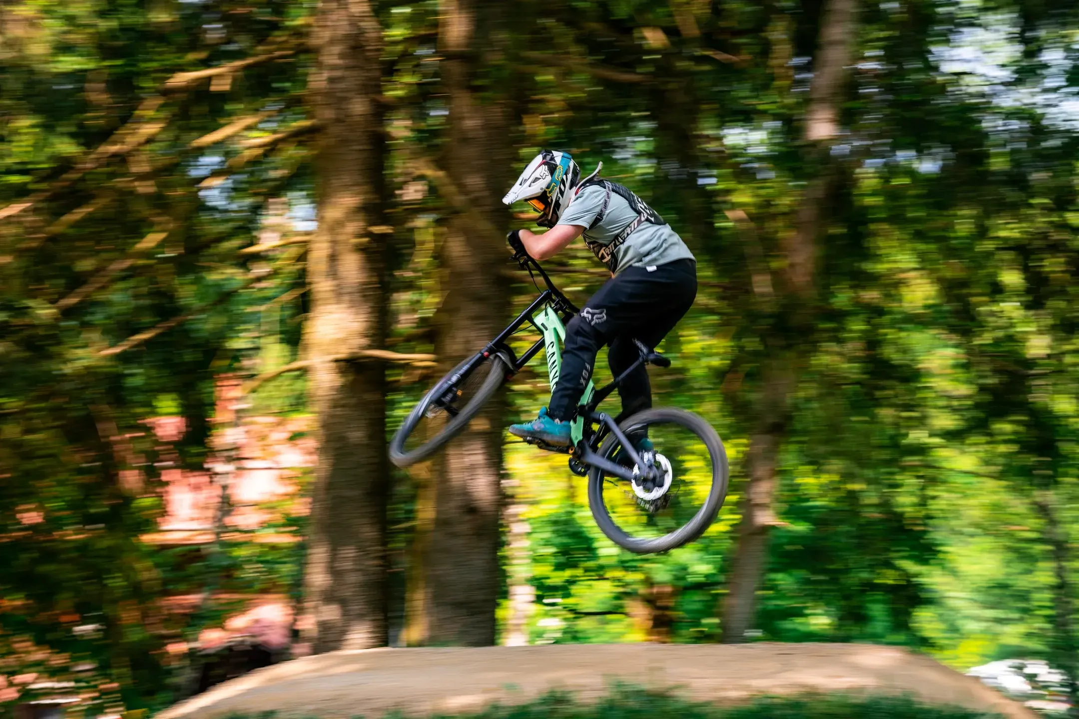 Junger Mountainbiker im Sprung über eine Erdrampe, Mitzieher mit verwischtem grünem Waldhintergrund