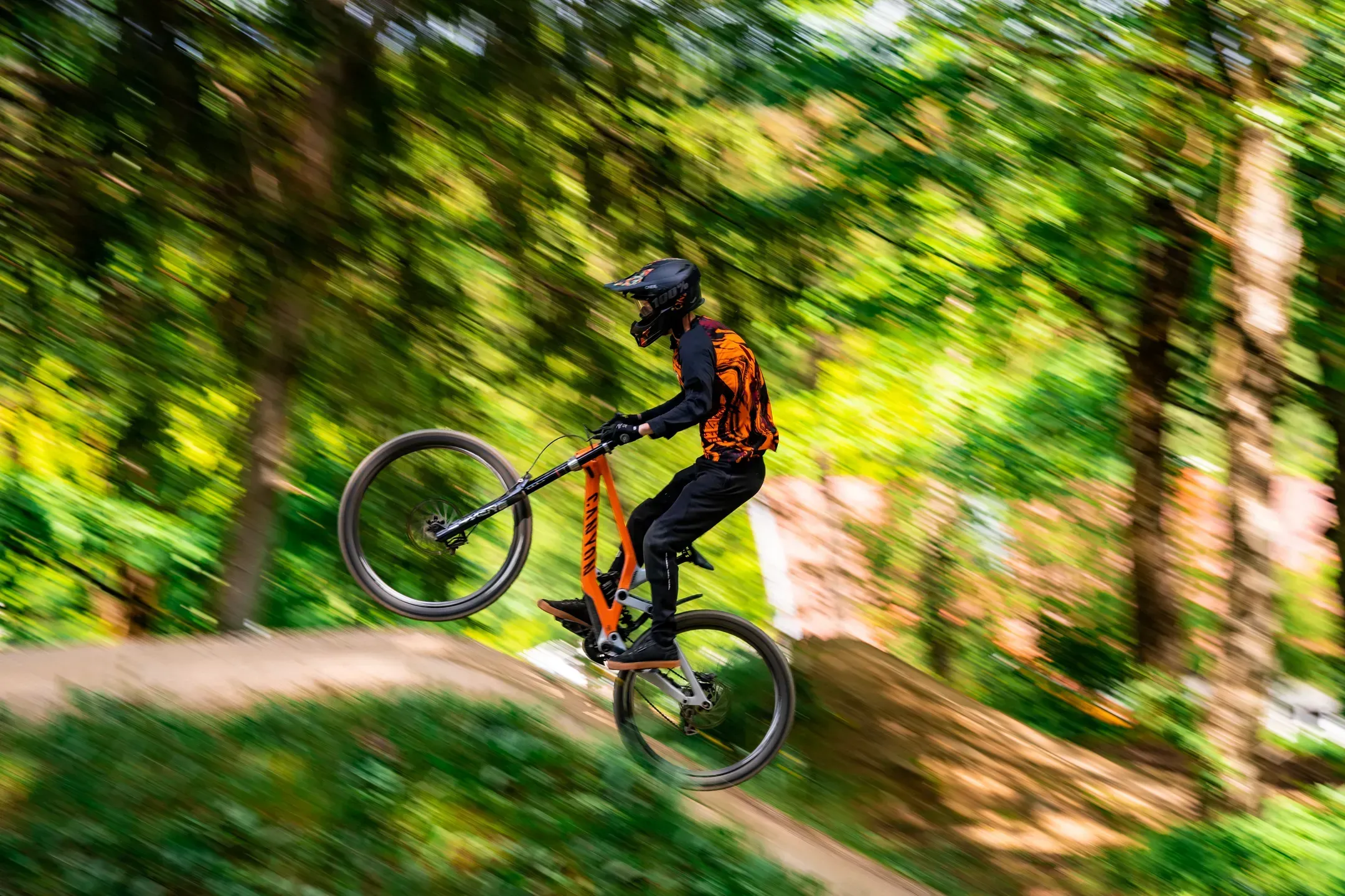 Mountainbiker mit orangem Rad und orangem Trikot im Sprung, Mitzieher vor verwischtem Laubwald