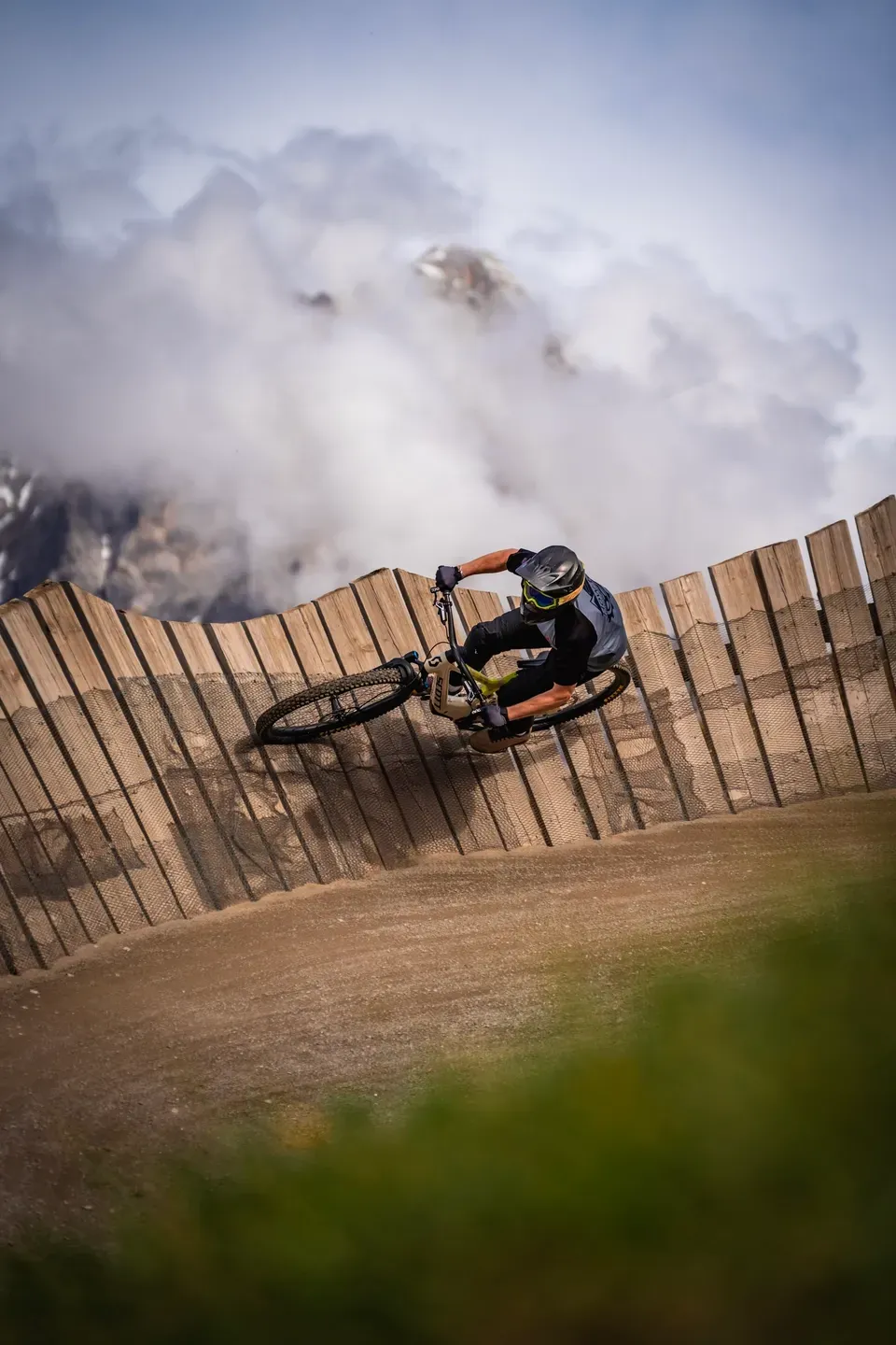 Mountainbiker in einer hölzernen Steilkurve, dahinter ziehen Wolken um einen felsigen Berggipfel