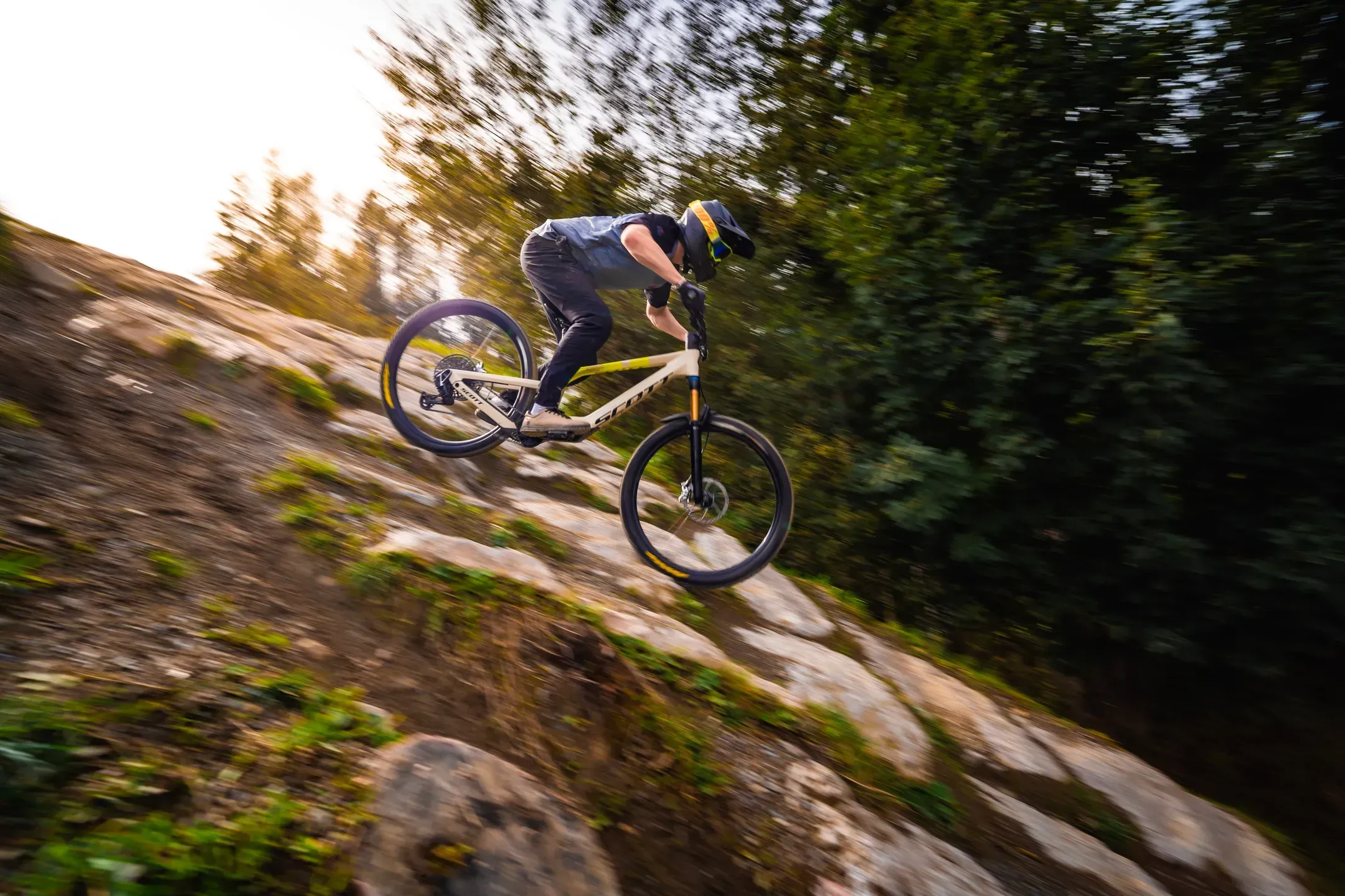 Mountainbiker fährt einen Felsabschnitt hinunter, Bewegungsunschärfe und warmes Gegenlicht am Waldrand