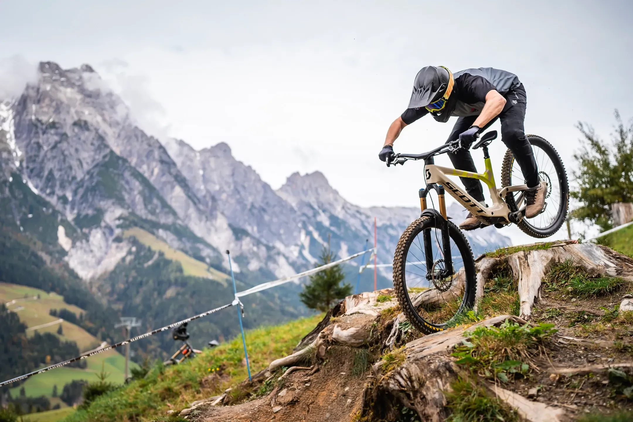 Mountainbiker springt über einen Baumstumpf auf einem Bergtrail, schroffe Felsberge im Hintergrund