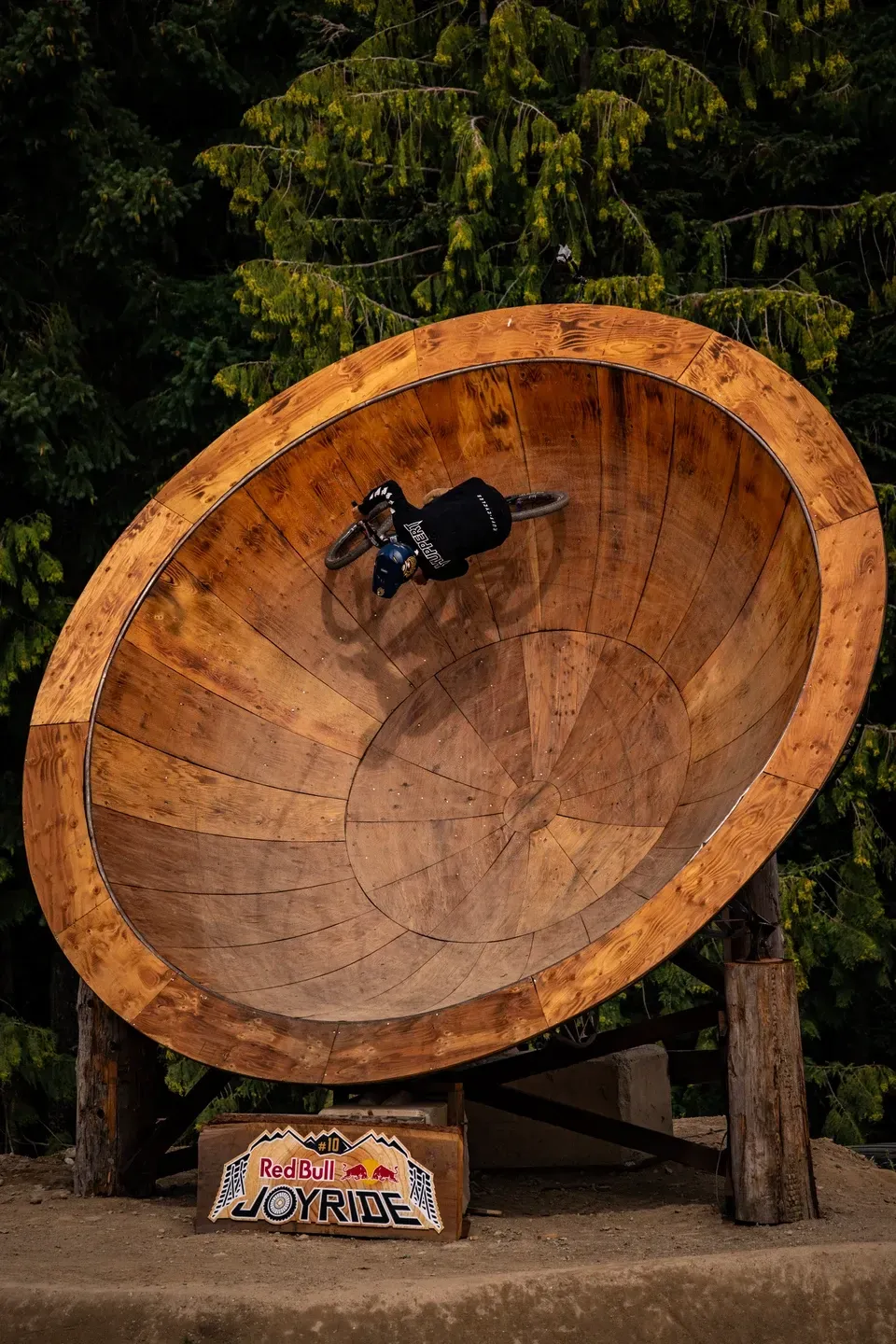 Mountainbiker fährt kopfüber an der Innenwand einer riesigen Holzschüssel, davor ein Red Bull Joyride Schild