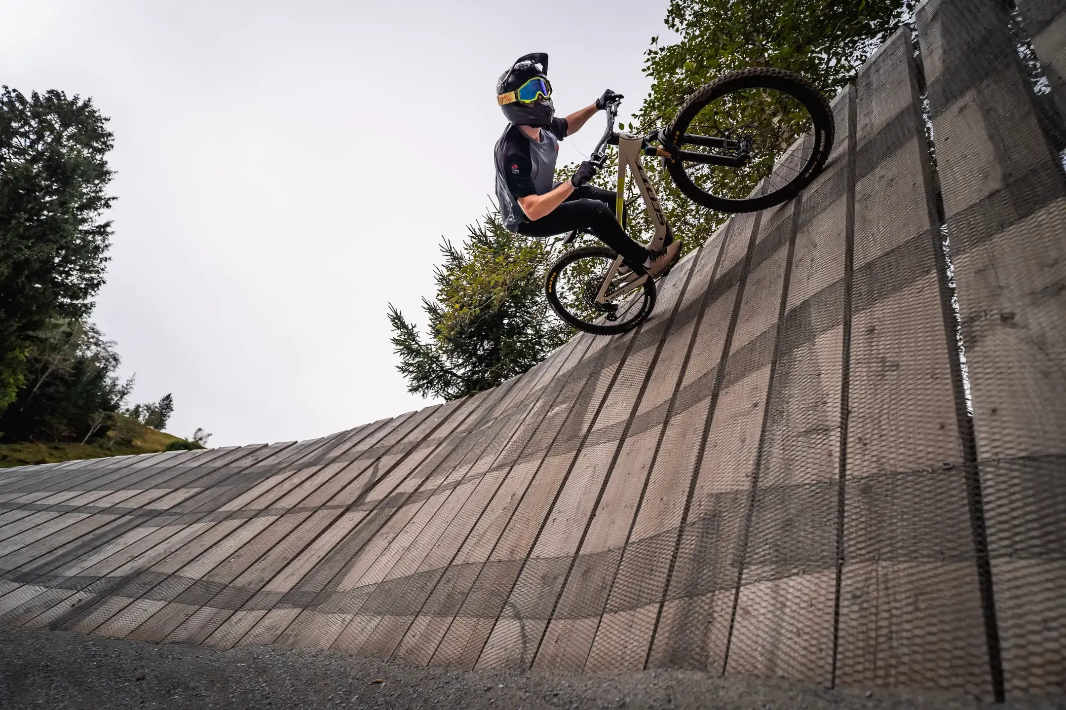 Mountainbiker auf dem Hinterrad an einer hohen Holzwand, Froschperspektive vor grauem Himmel