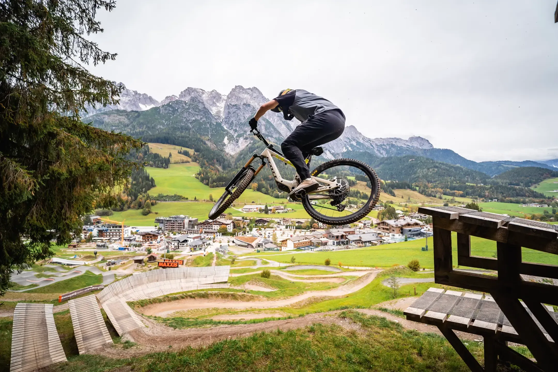 Mountainbiker springt von einer Holzrampe, unten Bikepark mit Holzkurven, ein Dorf und Felsgipfel