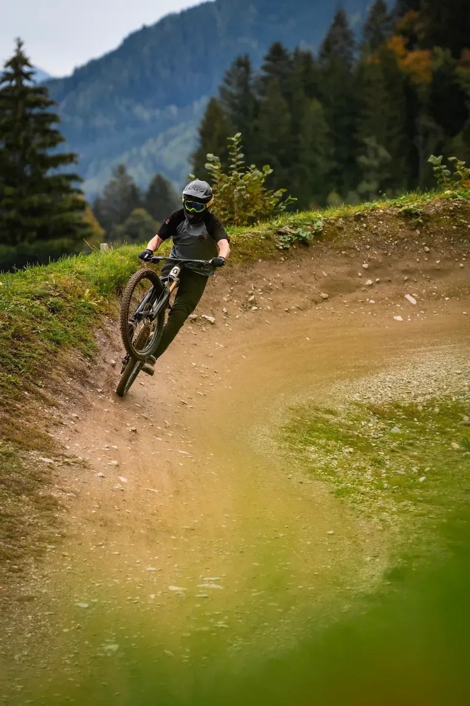Mountainbiker hebt in einer Erdkurve das Vorderrad an, Nadelwald und Berghang im Hintergrund