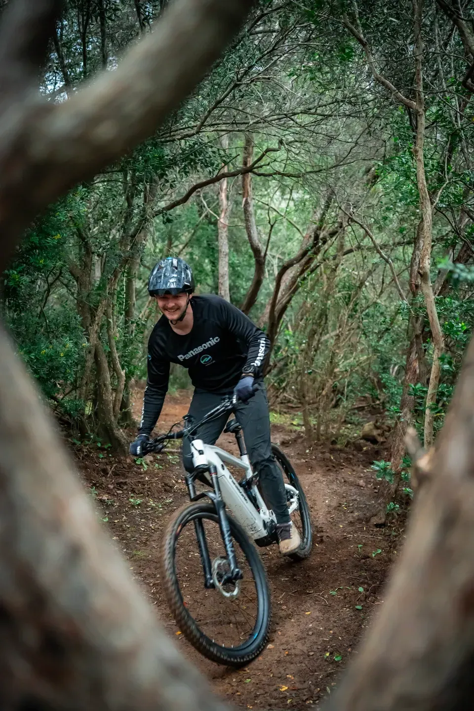 Lächelnder Mountainbiker mit weißem E-Bike auf einem Waldtrail, gerahmt von knorrigen Ästen