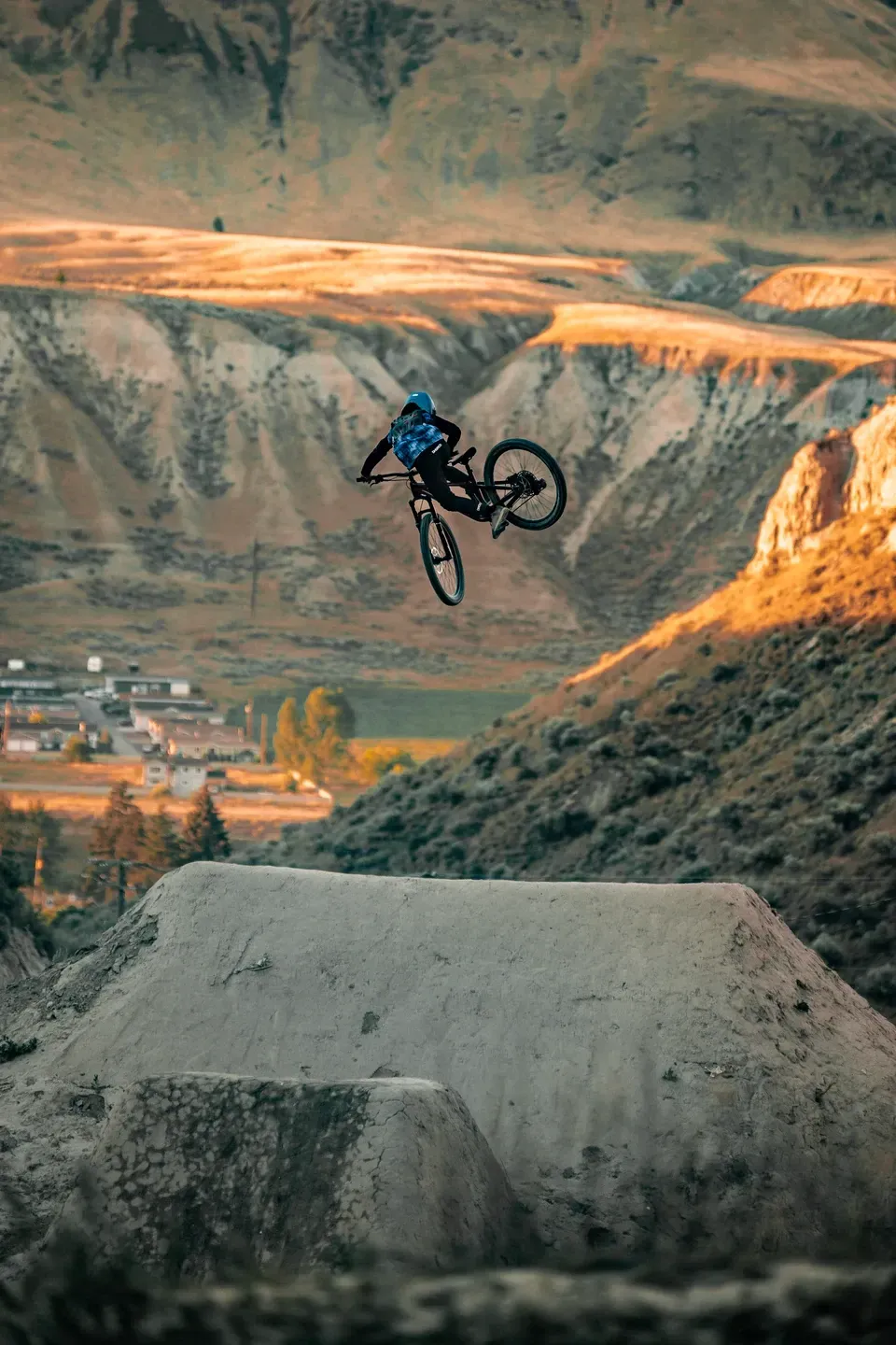 Mountainbiker im Whip über einem Erdsprung vor Hügeln im warmen Abendlicht, Häuser im Tal
