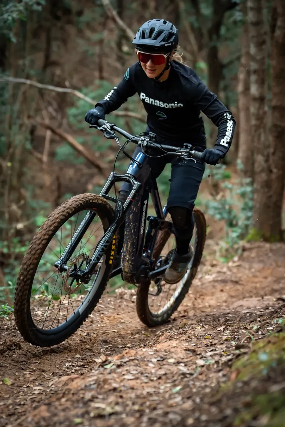Mountainbikerin mit Helm und Sportbrille fährt über einen laubbedeckten Waldtrail, Nahaufnahme von vorne