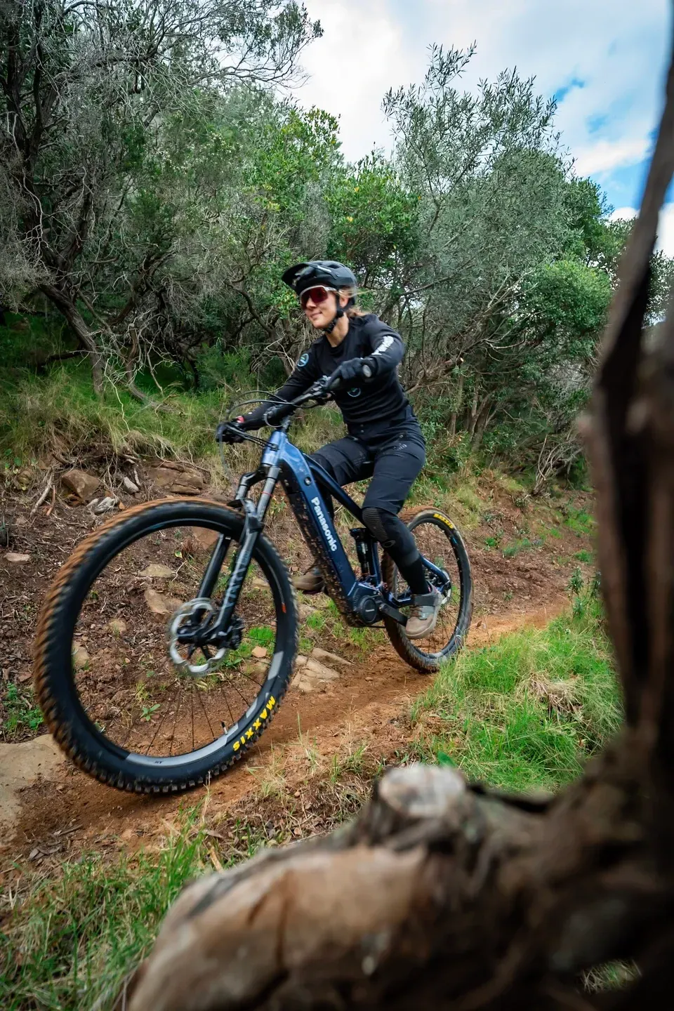 Mountainbikerin mit blauem E-Mountainbike auf einem Trail zwischen Buschwerk, Baumstamm im Vordergrund
