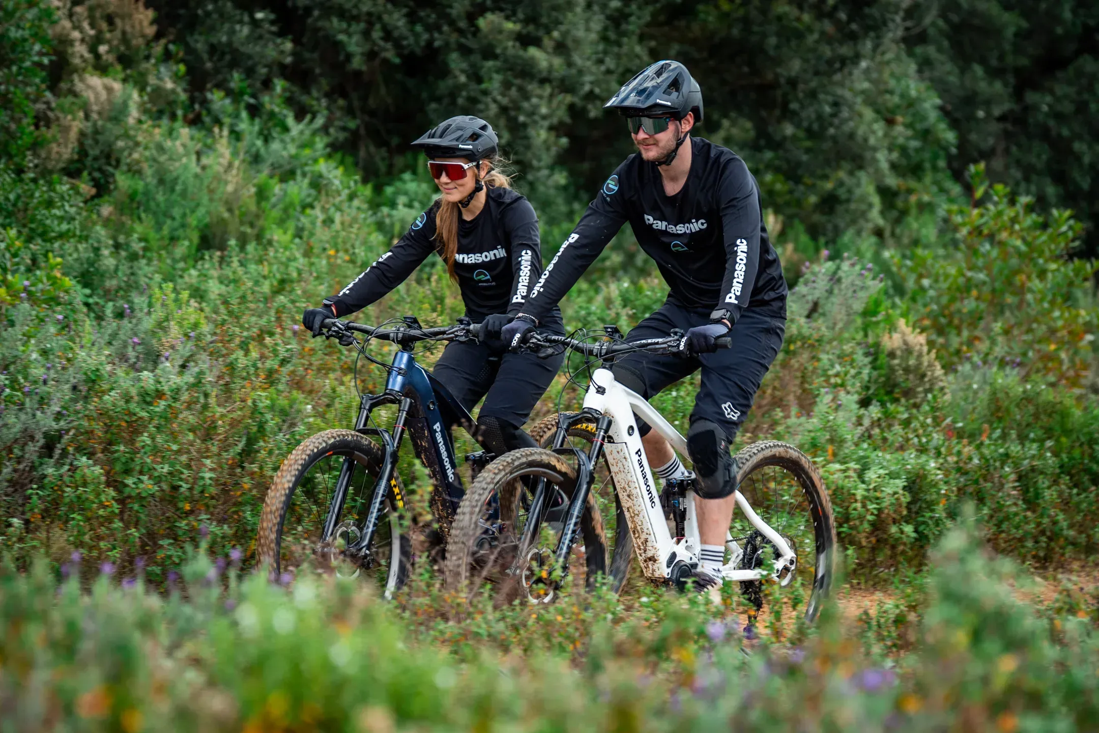 Frau und Mann fahren nebeneinander mit E-Mountainbikes durch blühendes Buschland, beide in Panasonic Trikots