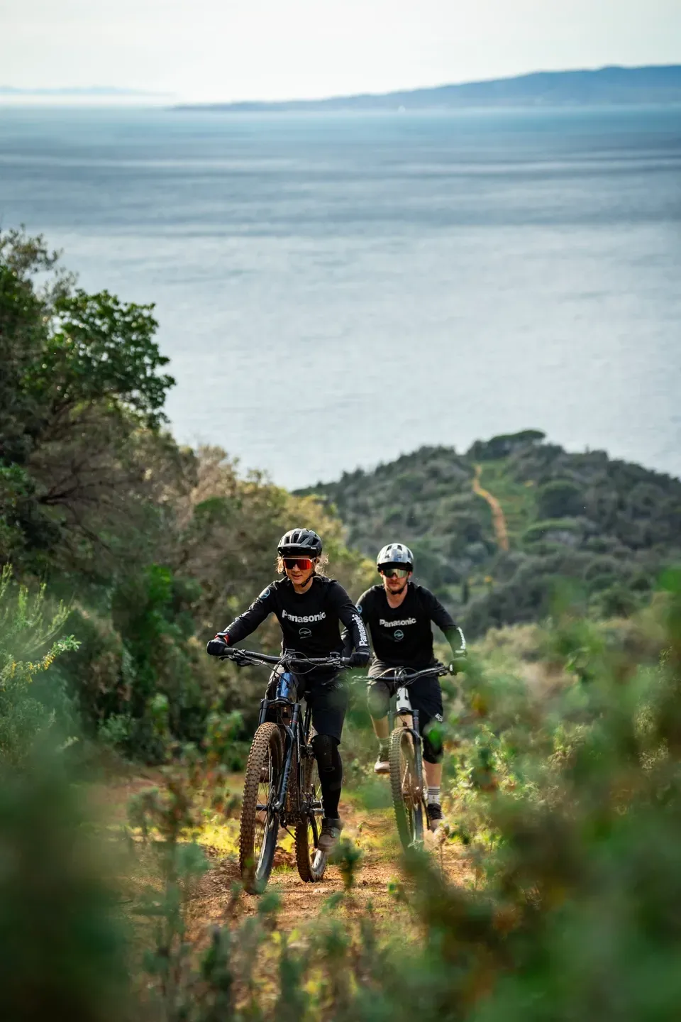 Zwei Mountainbiker fahren einen Trail durch Buschwerk hinauf, dahinter das Meer und eine Küstenlinie