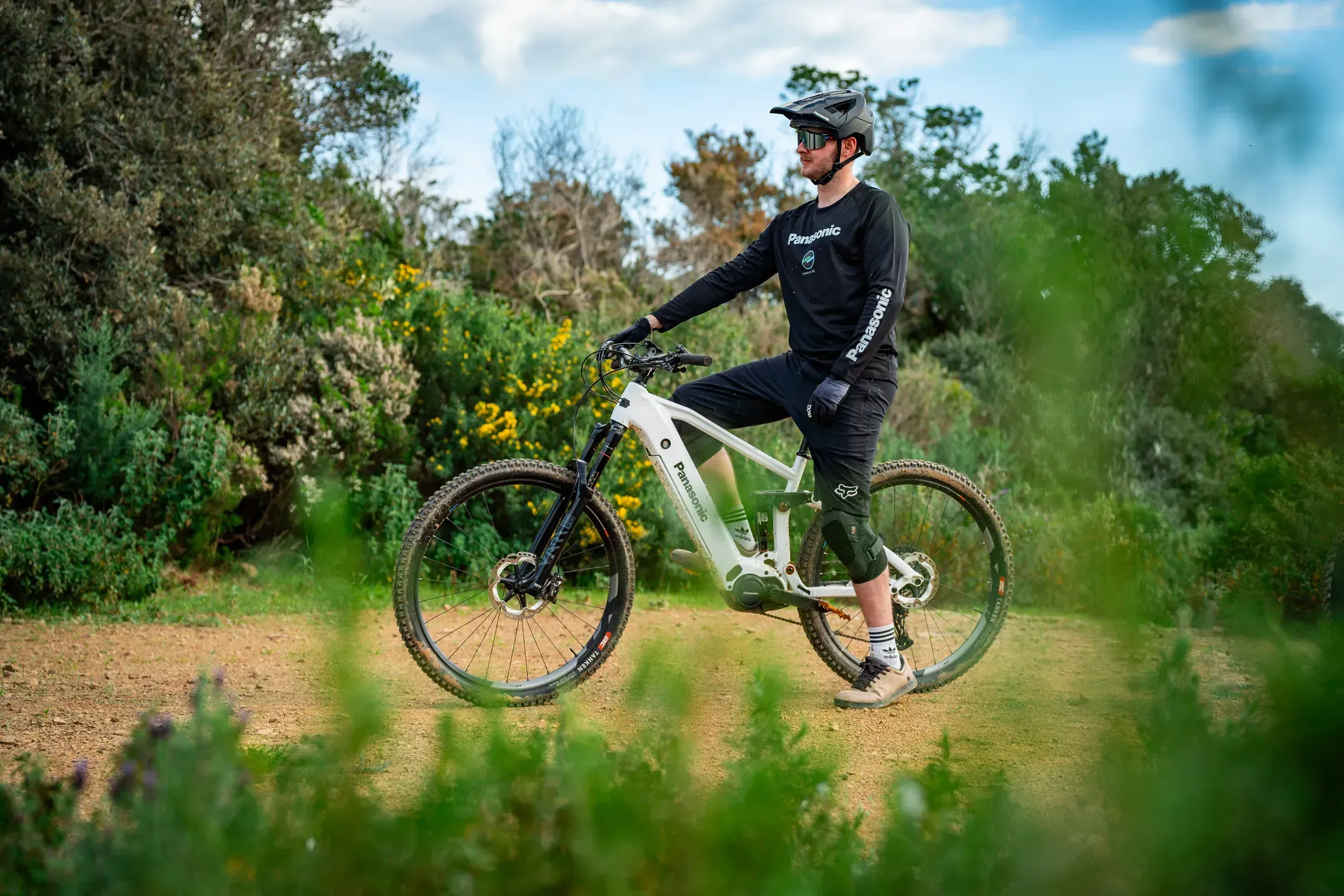 Mountainbiker im Panasonic Trikot steht mit weißem E-Mountainbike auf einem Schotterweg zwischen Büschen