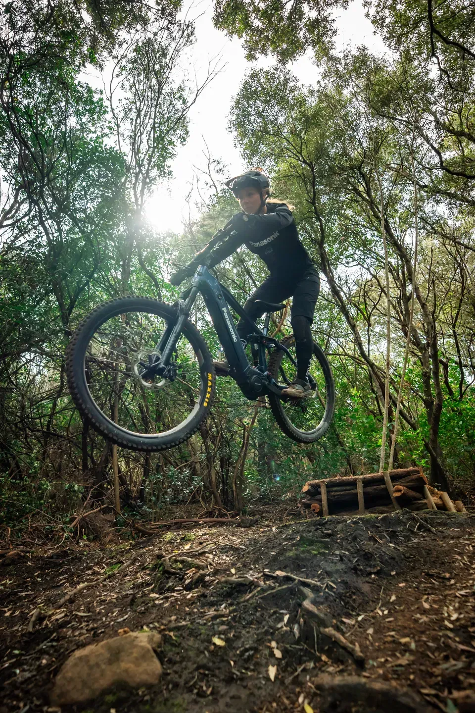Mountainbikerin springt mit dem E-Mountainbike über eine Erdkante im Wald, Gegenlicht durch die Bäume