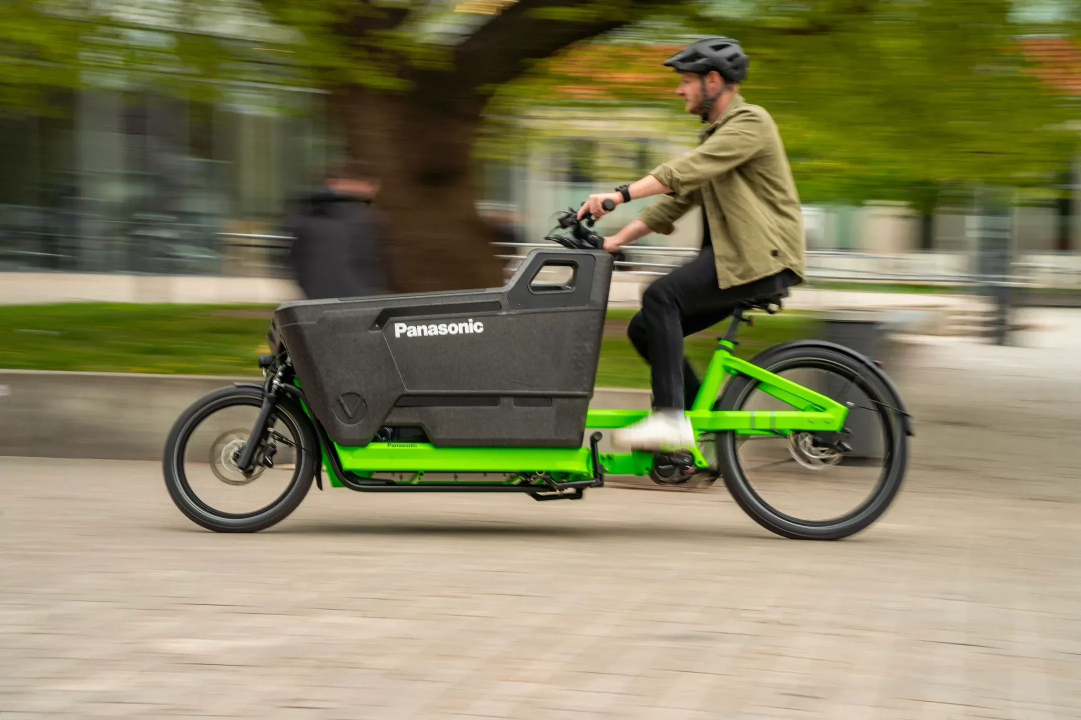 Mann fährt ein grünes Lastenrad mit schwarzer Panasonic Transportbox über einen gepflasterten Platz