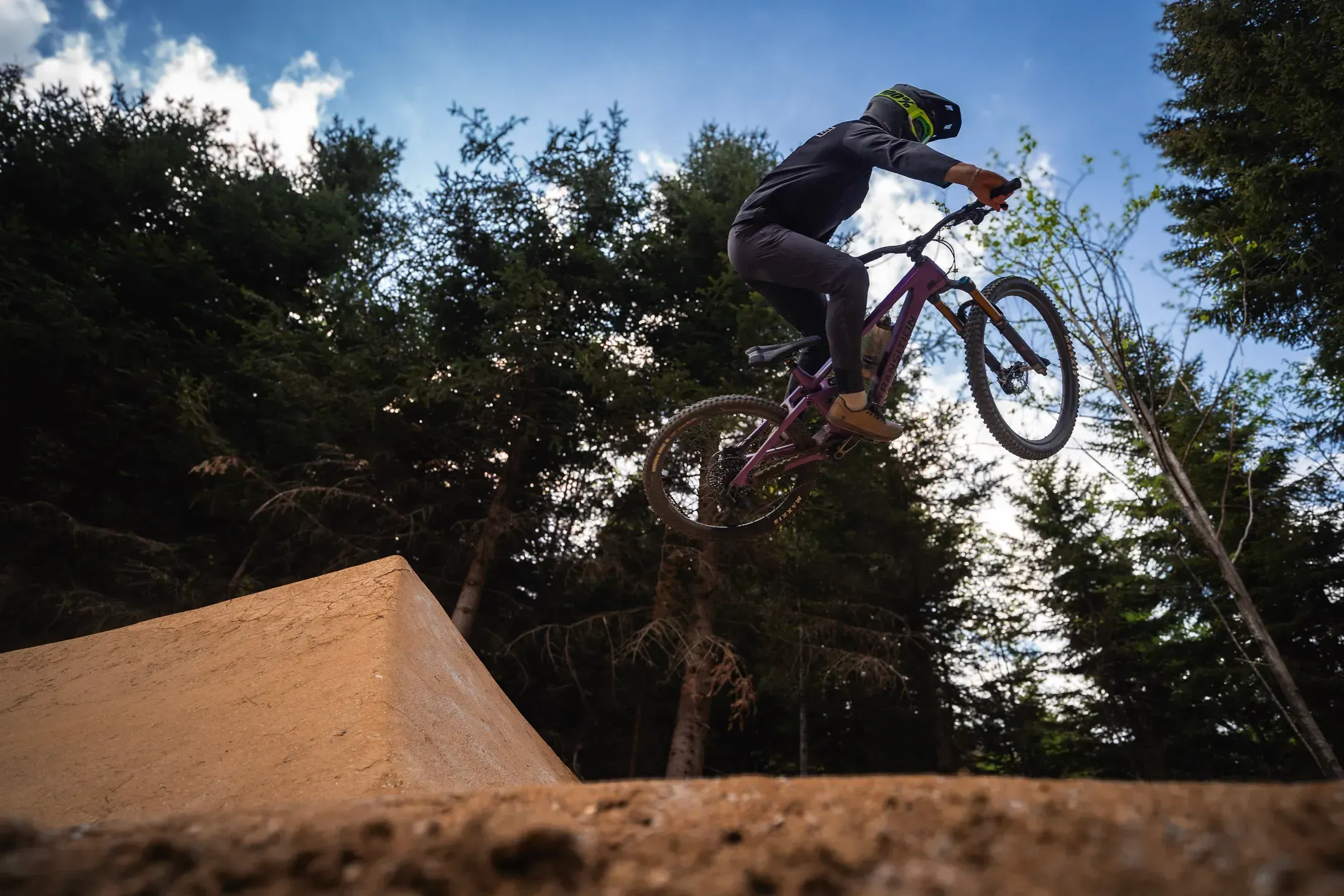 Mountainbiker springt aus der Froschperspektive über eine Lehmrampe, dahinter Nadelbäume und blauer Himmel