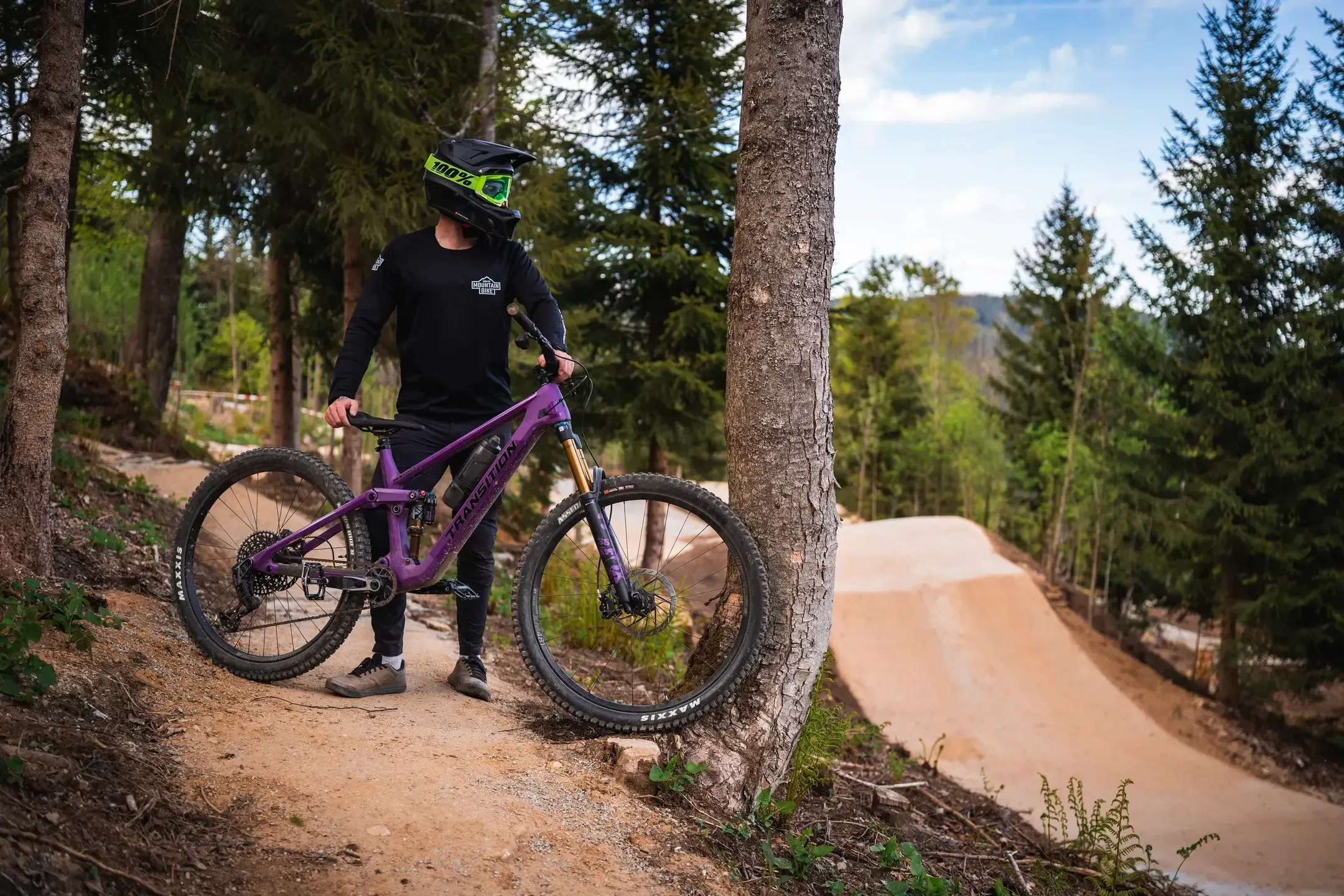 Mountainbiker mit Fullface-Helm steht mit lila Bike an einem Baum neben einer geschwungenen Lehmstrecke im Wald