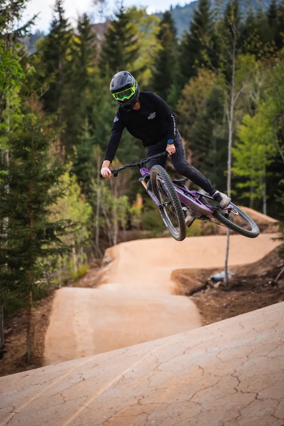 Mountainbiker im Sprung über eine Lehmwelle im Bikepark, im Hintergrund bewaldete Hänge und weitere Kurven