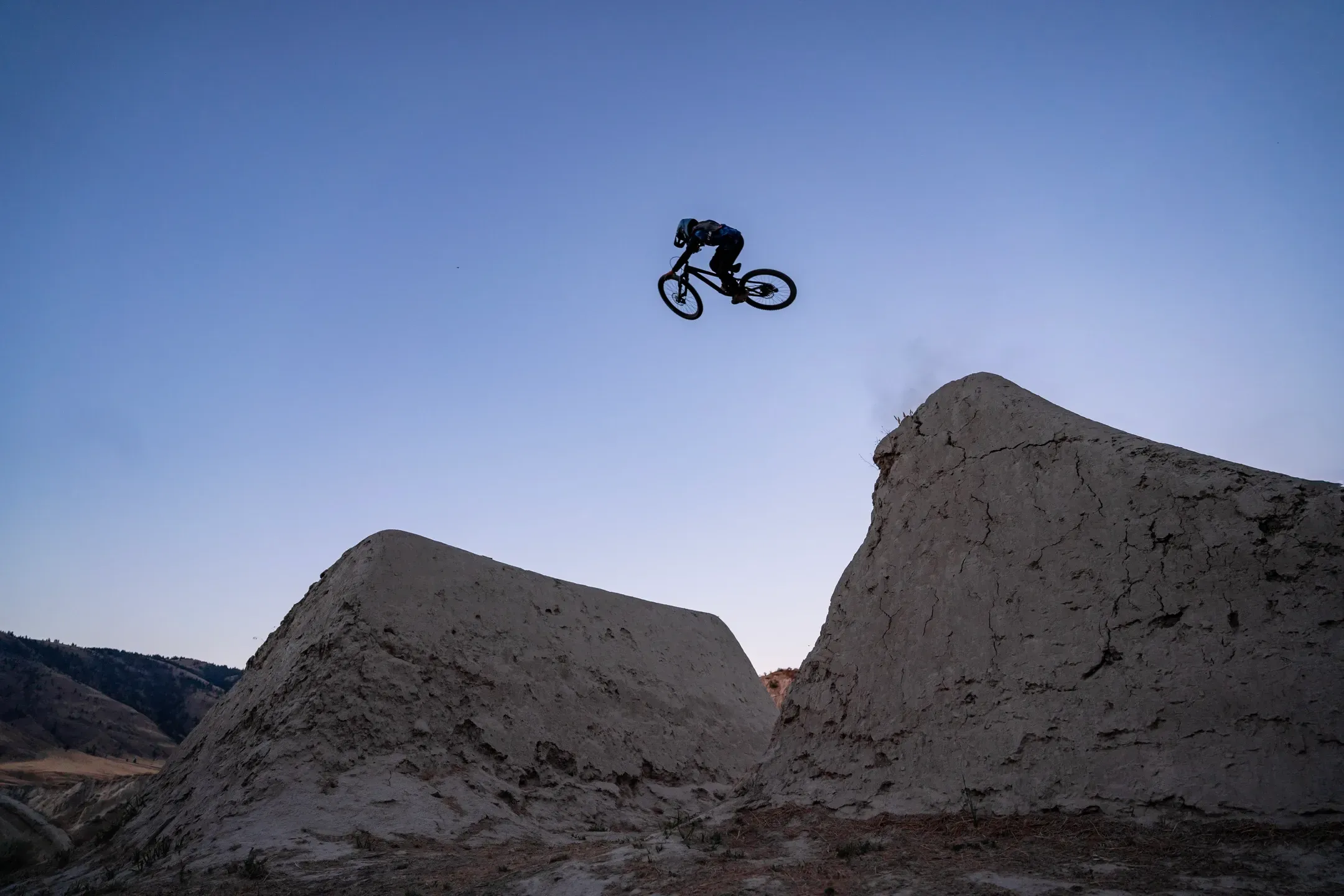 Silhouette eines Mountainbikers fliegt zwischen zwei steilen Lehmschanzen vor blauem Abendhimmel