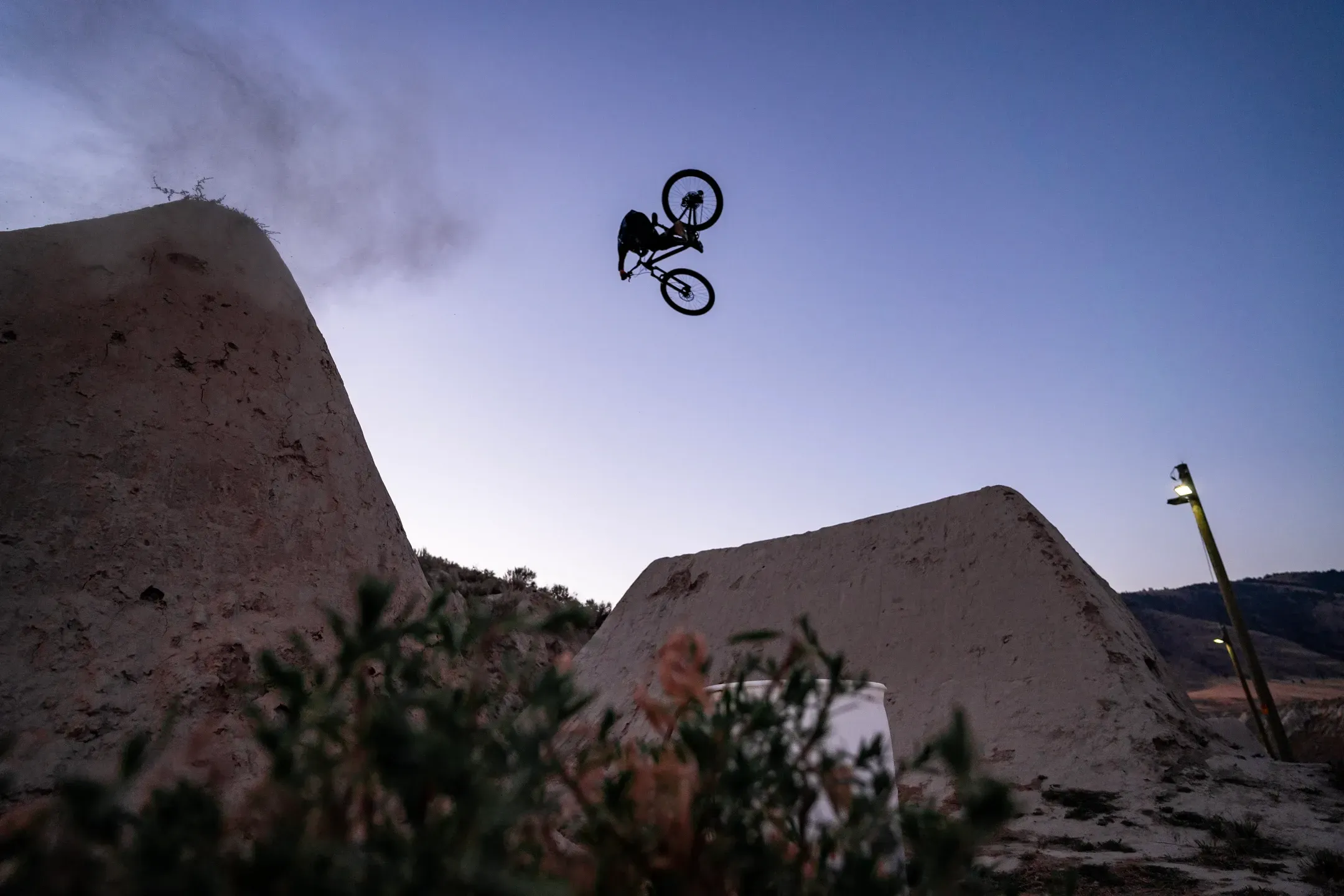 Mountainbiker kopfüber im Flip über einer Lehmschanze in der Dämmerung, Staubwolke und Laterne im Hintergrund