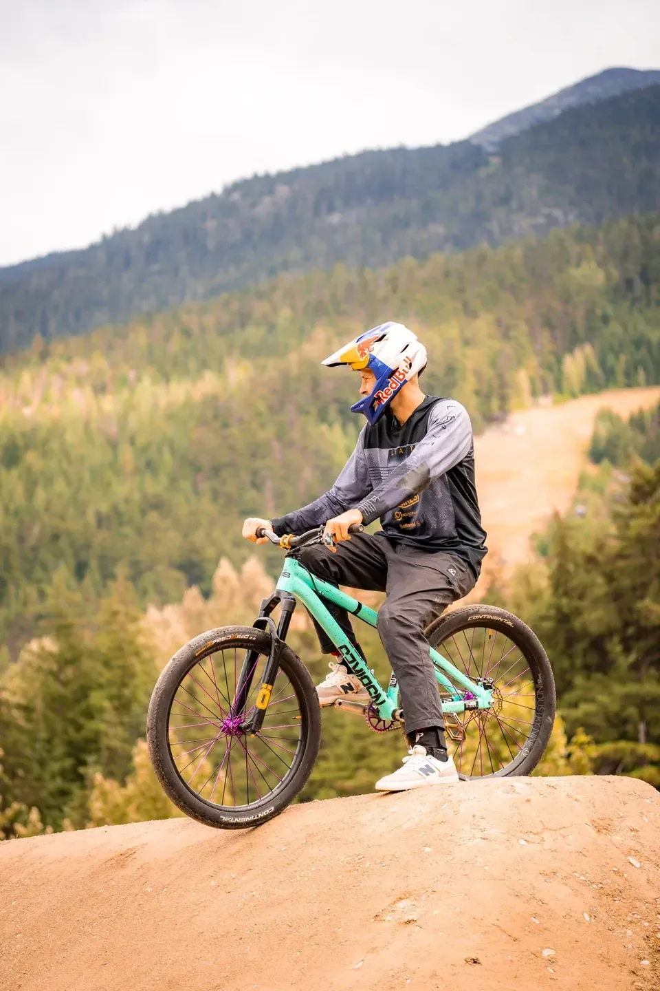 Dirtbiker mit buntem Helm steht auf einem Erdhügel und blickt über ein bewaldetes Bergtal