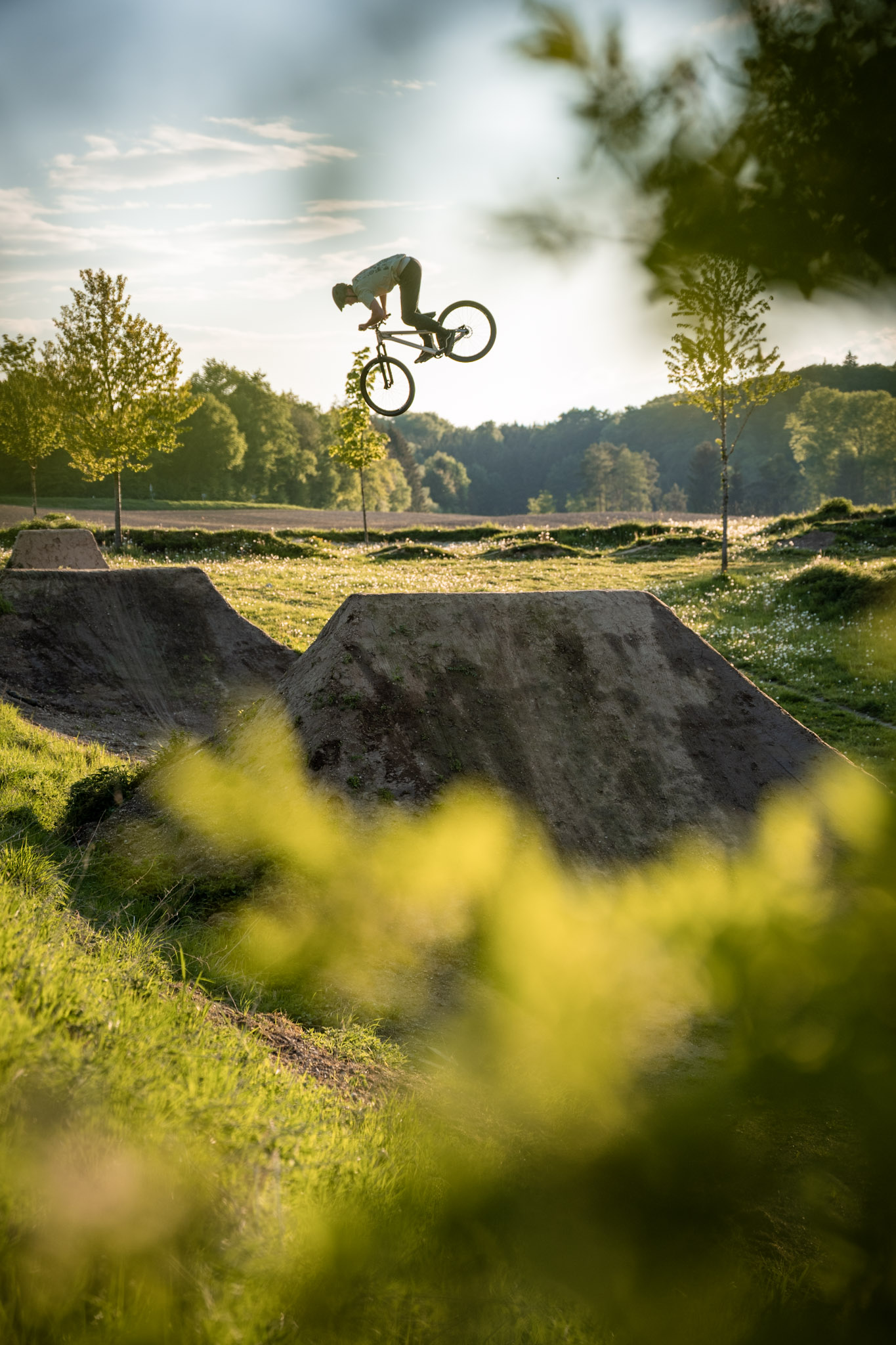 Dirtbiker streckt sich im Sprung über Erdschanzen auf einer Wiese im Abendlicht, Laub unscharf im Vordergrund