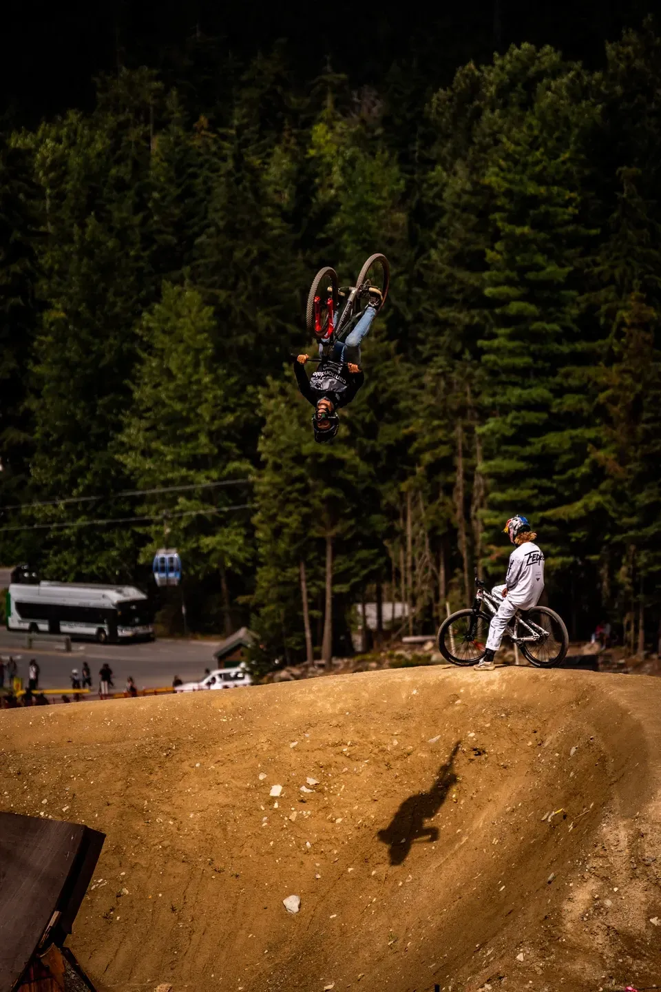 Dirtbiker im Backflip über einer Lehmschanze, ein zweiter Fahrer schaut zu, im Hintergrund eine Gondel