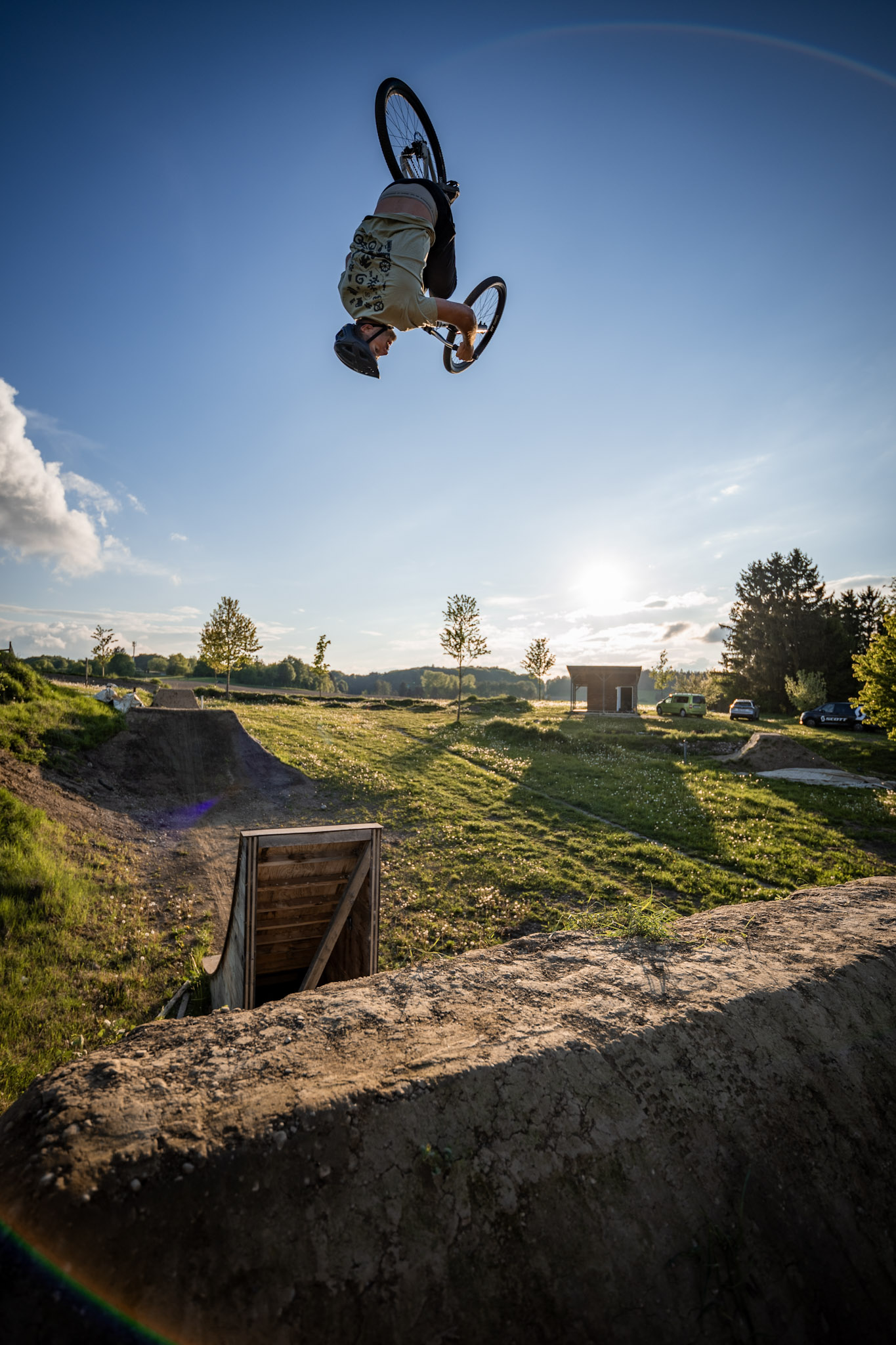 Dirtbiker kopfüber im Backflip vor blauem Himmel, darunter Erdschanze mit Holzrampe und tief stehende Sonne