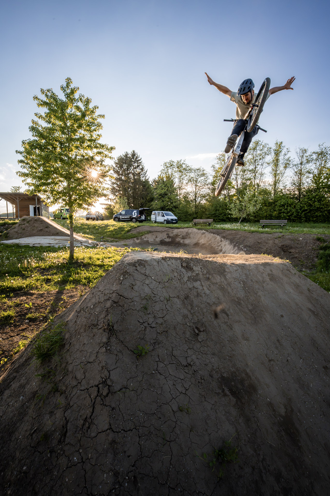 Dirtbiker breitet im Sprung beide Arme aus, unter ihm ein rissiger Erdhügel und Gegenlicht durch einen Baum