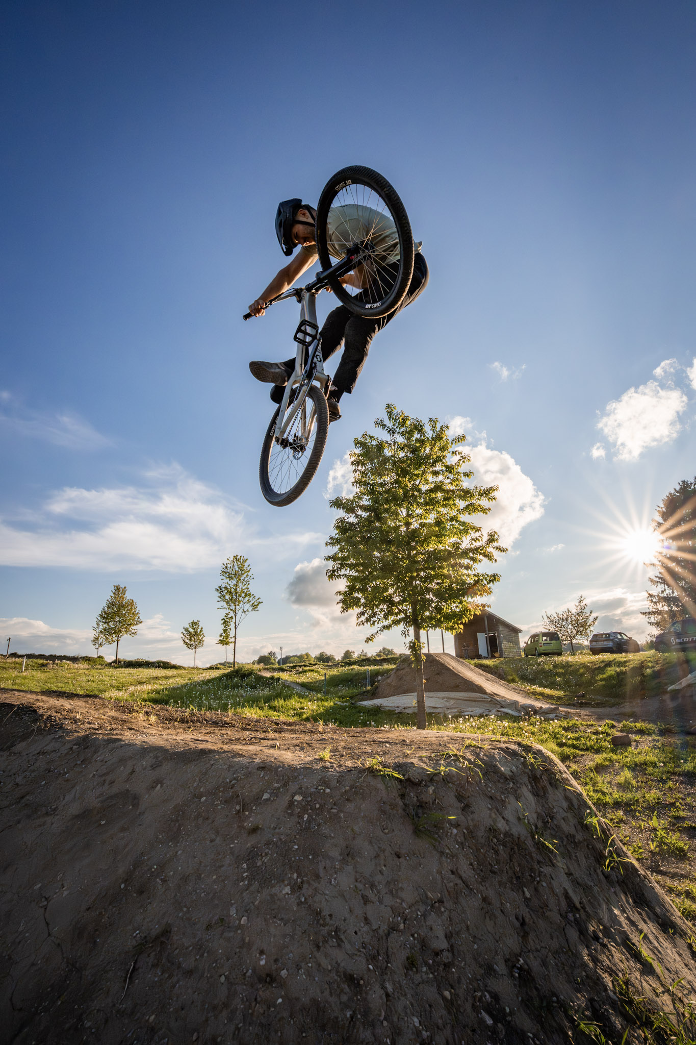 Dirtbiker im Sprung aus der Froschperspektive, Sonnenstern hinter einem jungen Baum und Wolken am Himmel