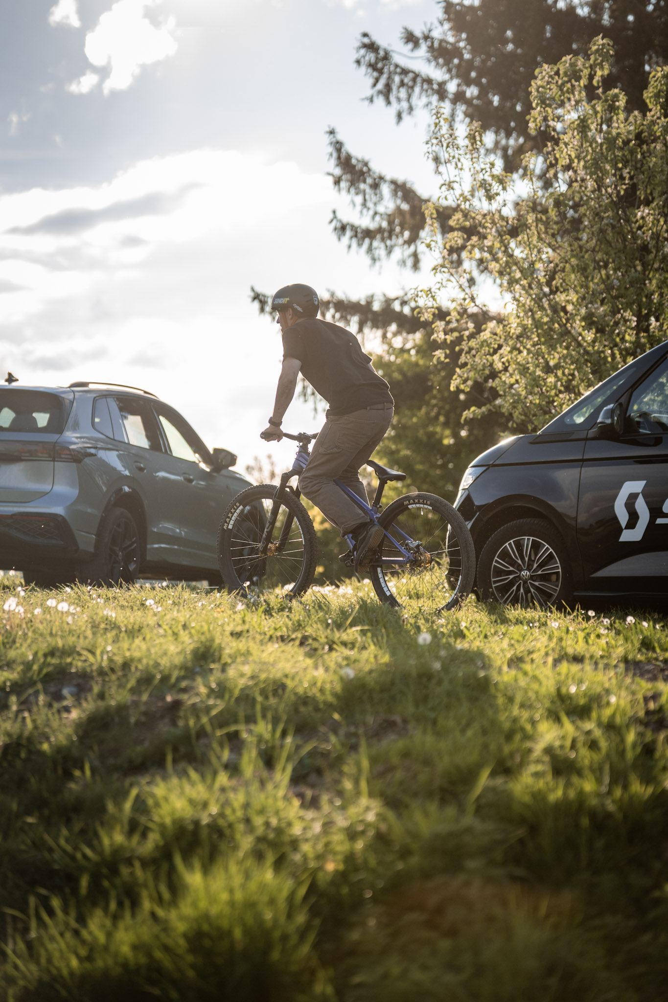 Mountainbiker rollt im warmen Gegenlicht über eine Wiese zwischen zwei geparkten Autos
