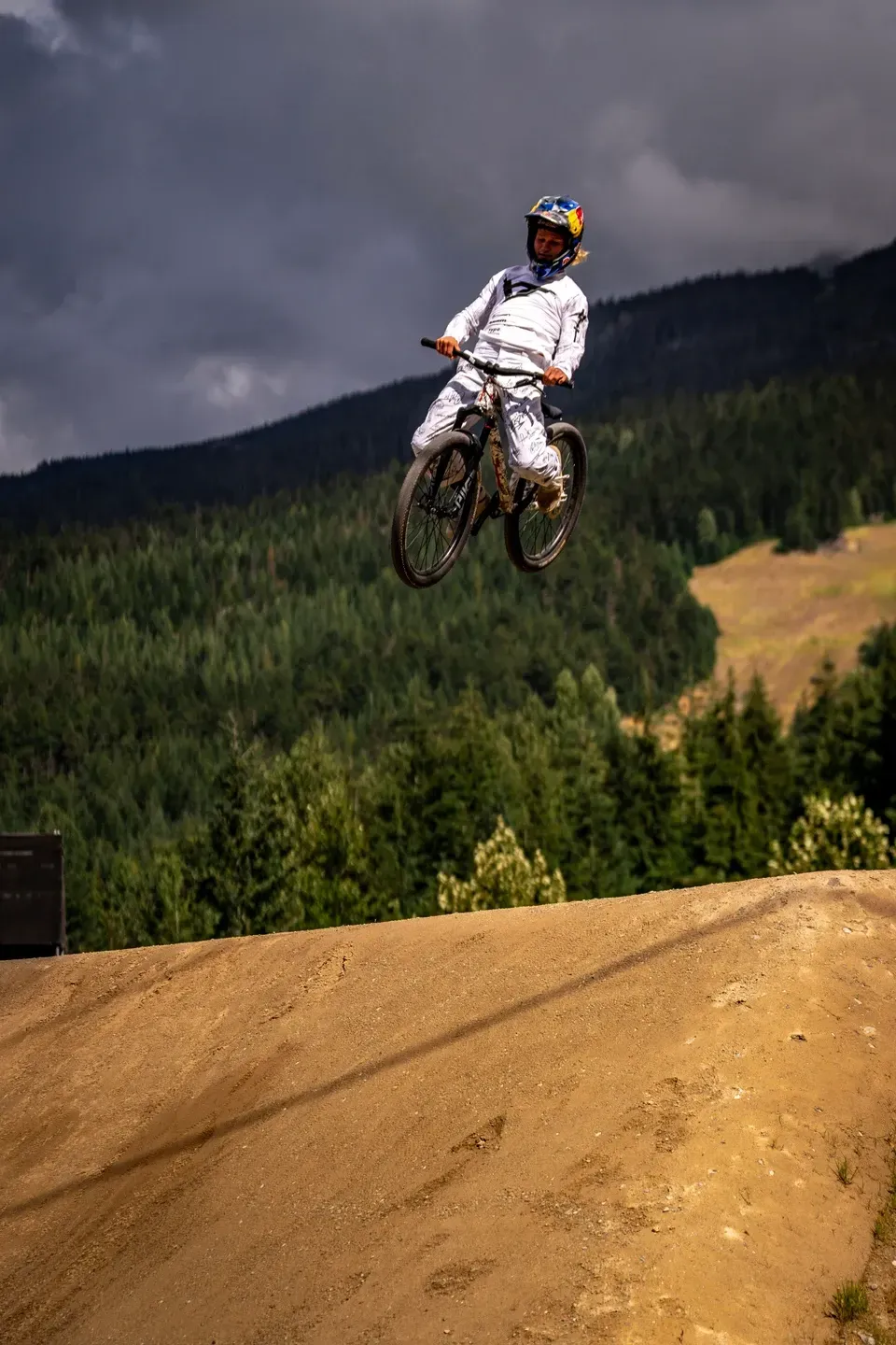 Mountainbiker in weißem Outfit springt über eine Lehmschanze, dahinter bewaldete Berge und dunkle Gewitterwolken