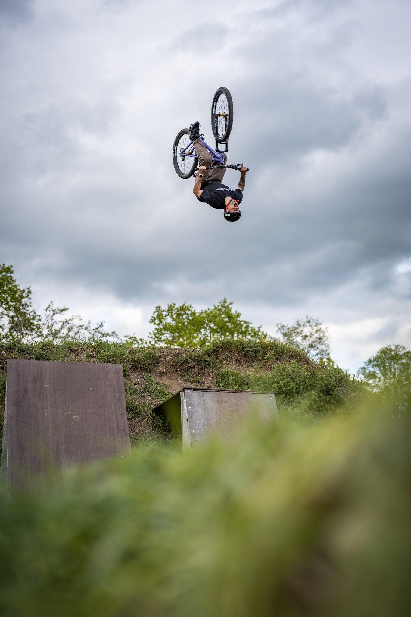 Dirtbiker kopfüber im Backflip unter grauem Wolkenhimmel, darunter zwei Holzrampen und unscharfes Gras