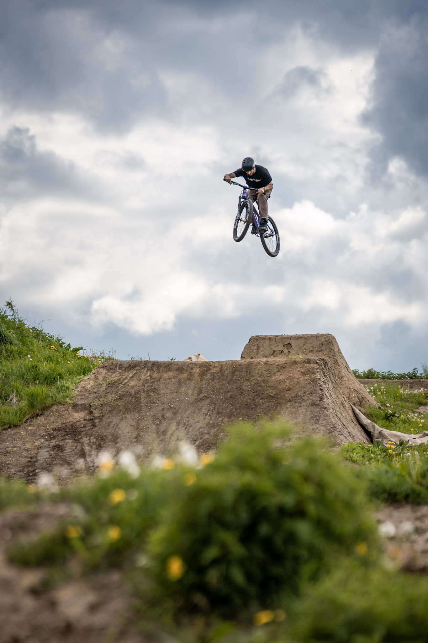Dirtbiker im Sprung vor dramatischen Wolken über einer Erdschanze, gelbe Blumen unscharf im Vordergrund