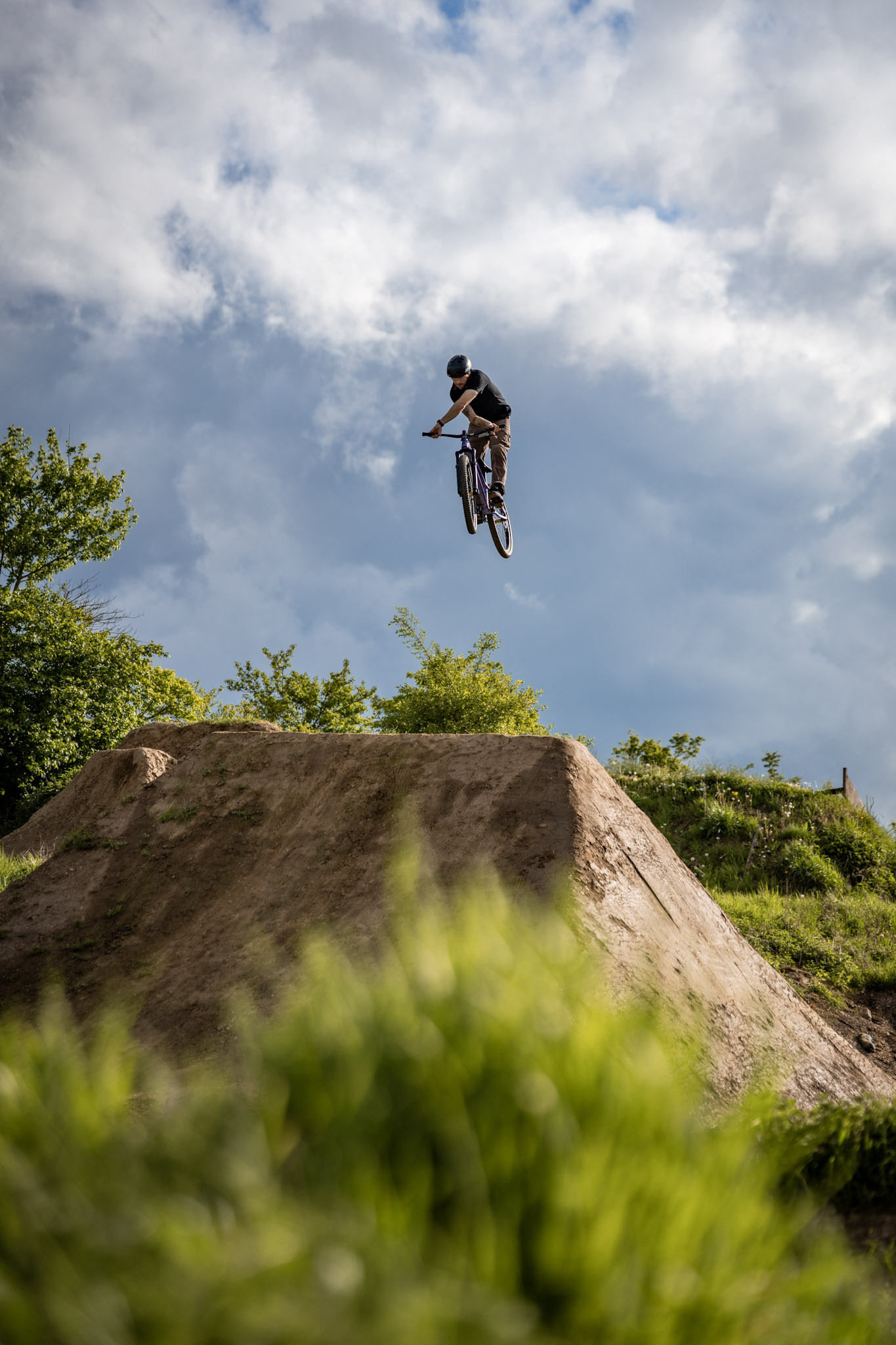Dirtbiker mit lila Bike im Tabletop über einer hohen Erdschanze, Wolken und grüne Büsche im Hintergrund