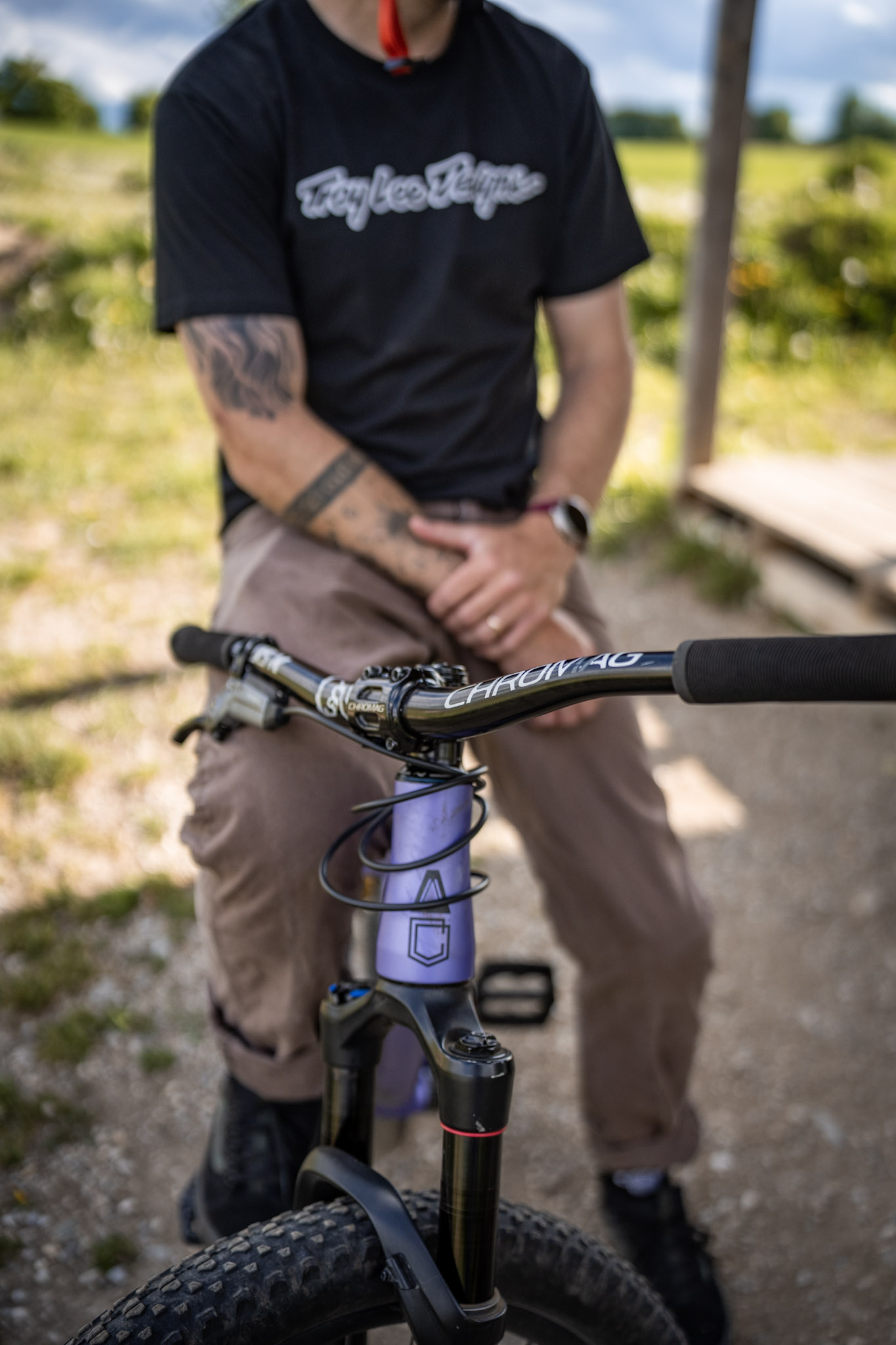 Sitzender Fahrer mit tätowiertem Arm hinter dem Lenker seines lila Dirtbikes, Gabel scharf im Vordergrund